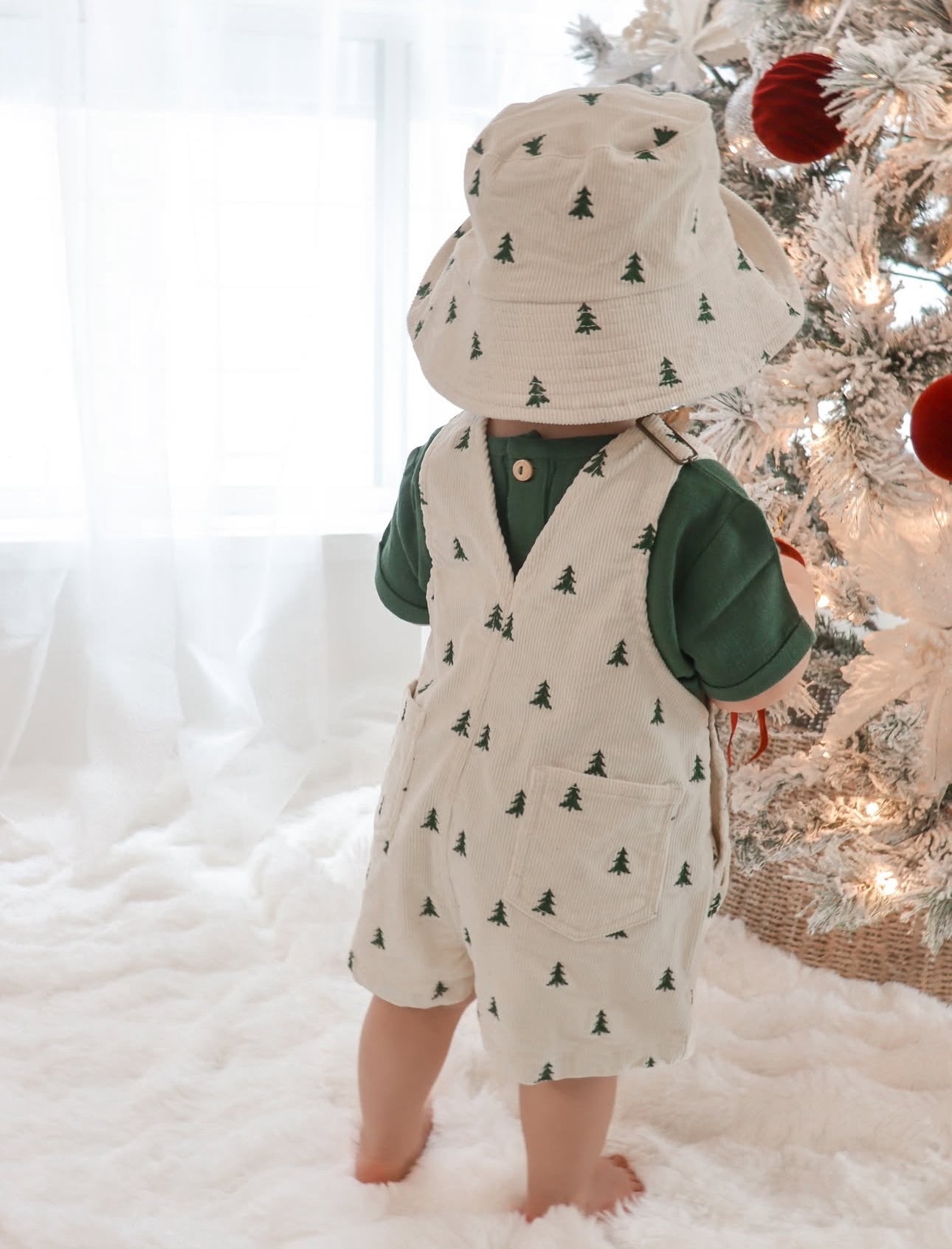 Toddler in Christmas tree print corduroy overalls, a green tee and matching bucket hat beside a flocked tree