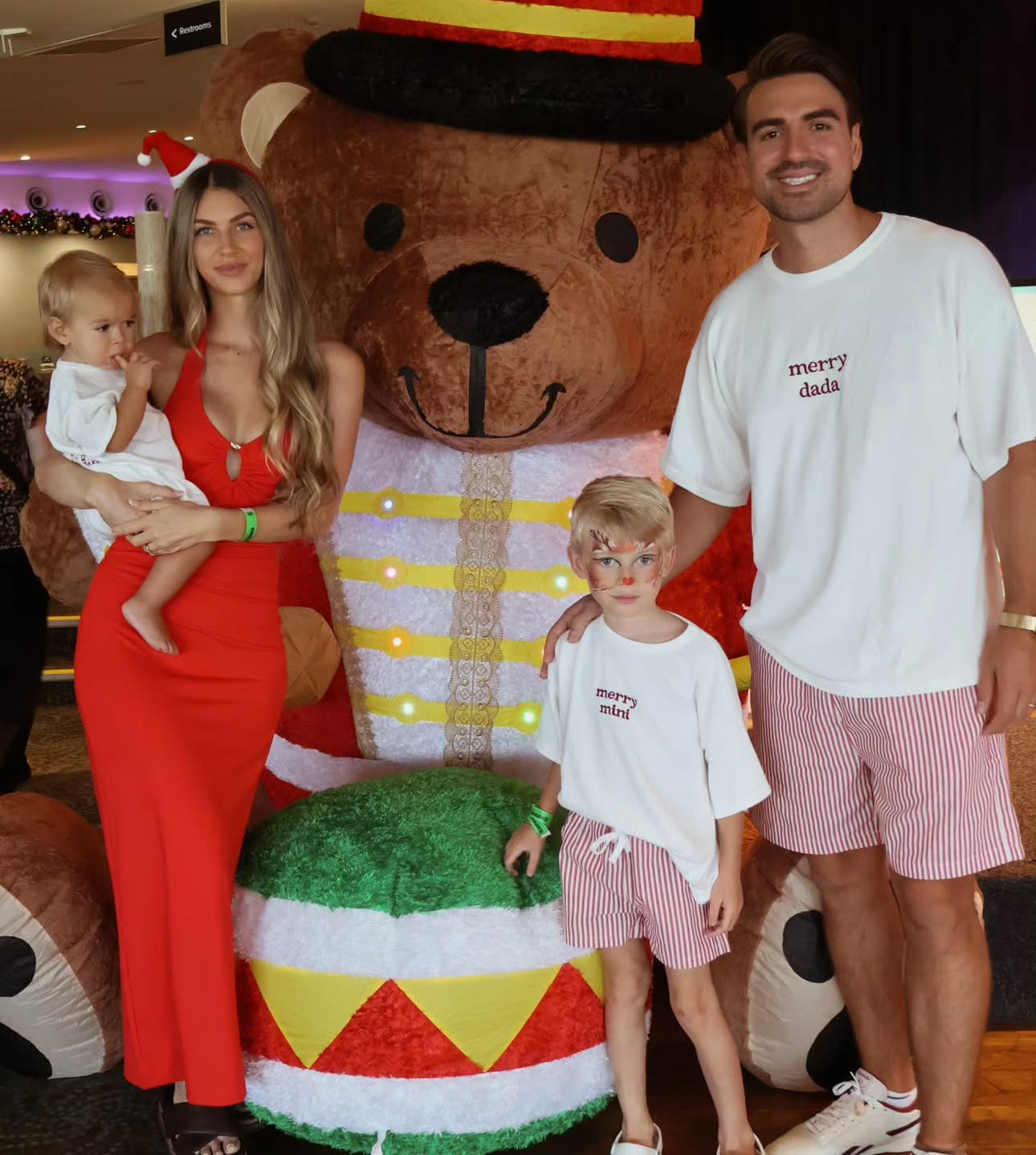 Family in matching merry mini and merry dada tees with red stripe shorts at a Christmas display