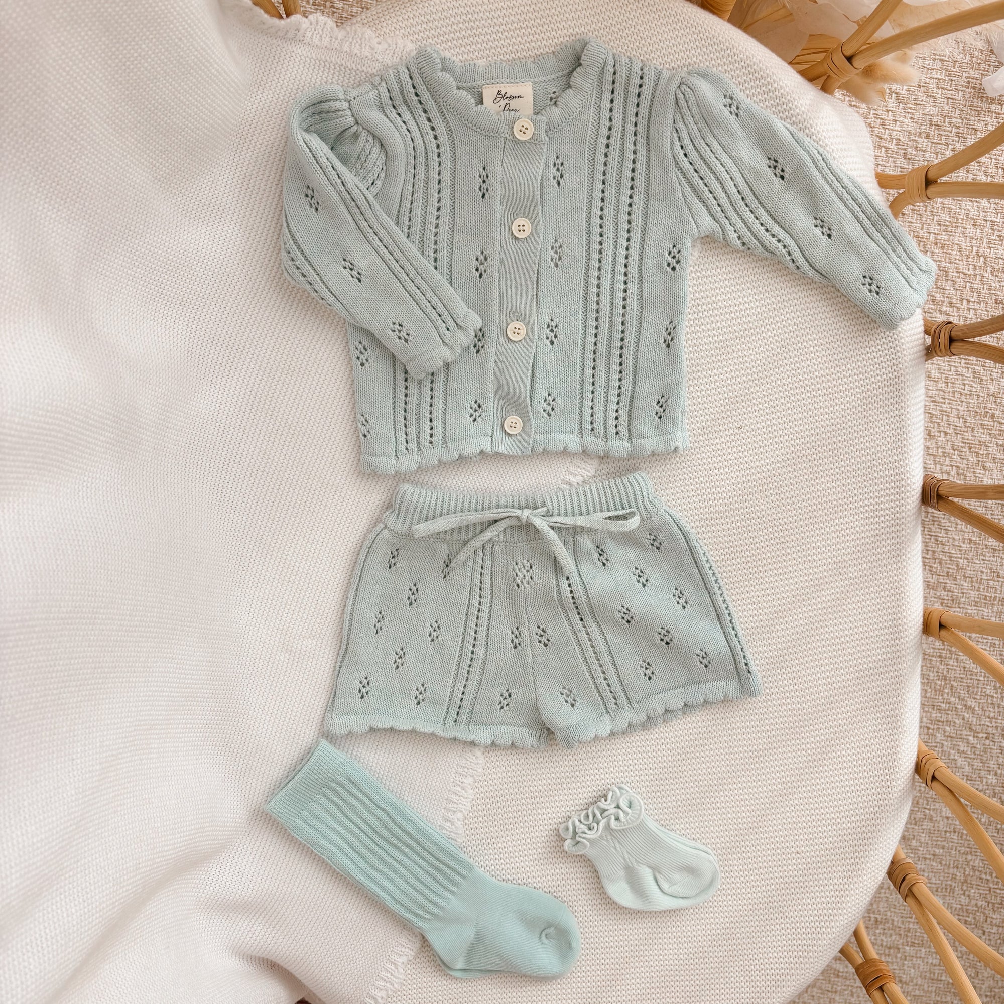 Blue pointelle knit baby cardigan and shorts set with matching socks, flat lay on a cream blanket