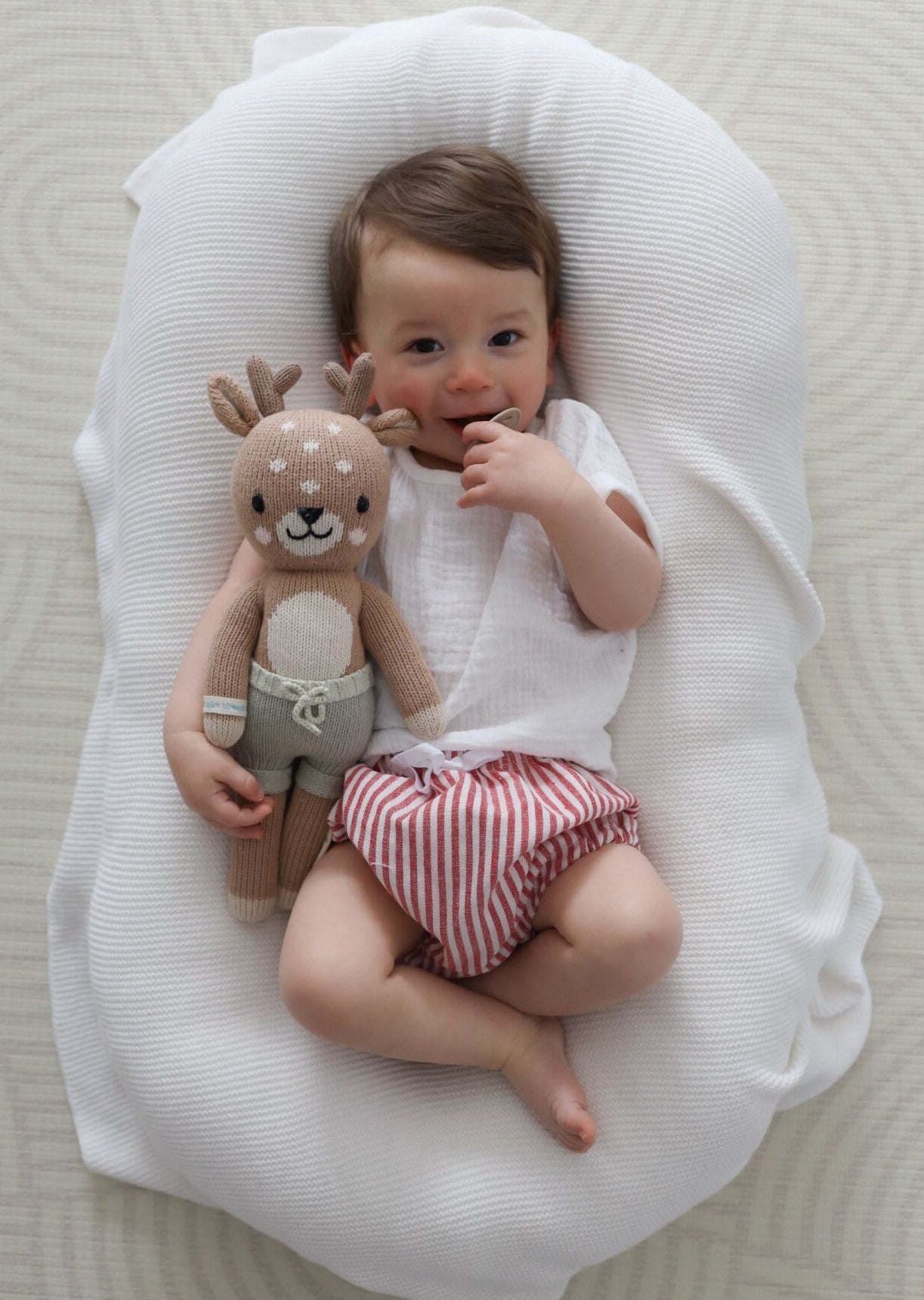 Baby in a white top and red stripe bloomers lying on a knit blanket beside a knitted reindeer toy
