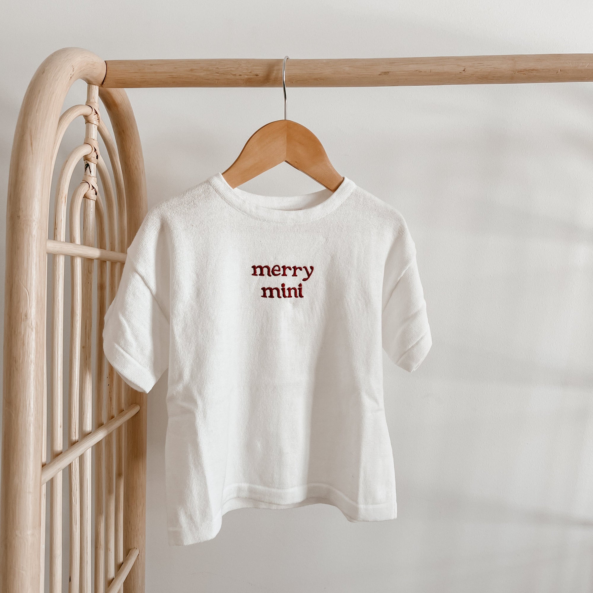 White kids tee embroidered with merry mini in red, hanging on a rattan clothing rack