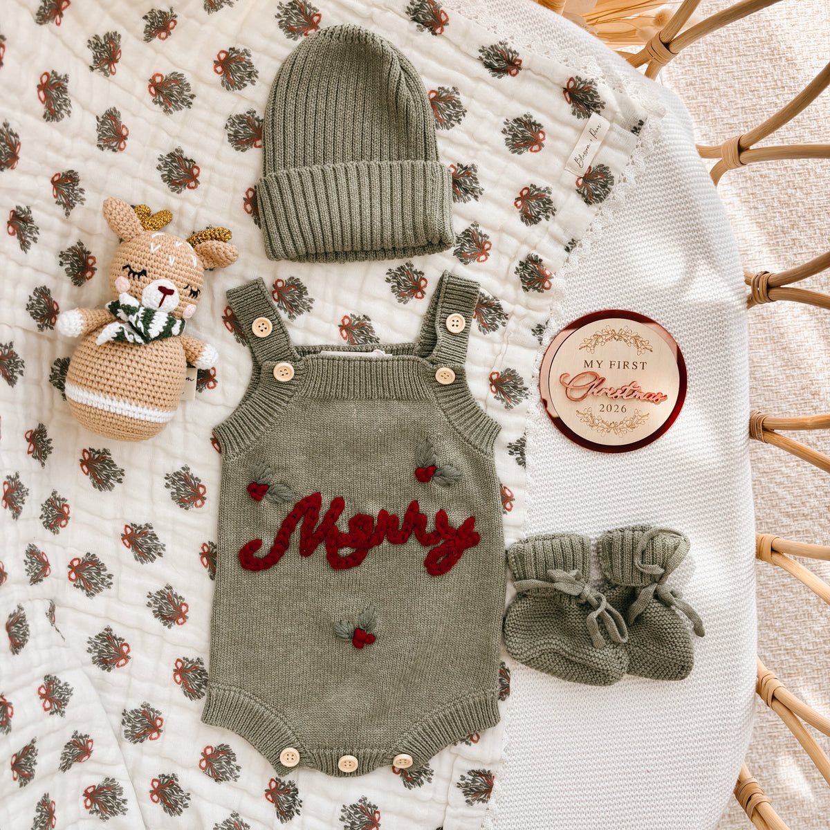 Sage green Merry knit overall romper flat lay with a matching beanie, booties and crochet reindeer rattle beside a My First Christmas 2026 keepsake disc