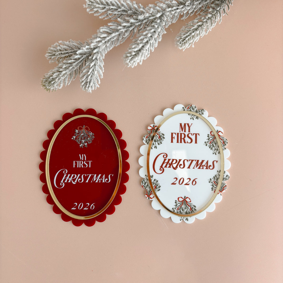 Two oval Christmas ornament-style plaques on a peach background, one red with gold trim and one white with floral trim, both reading 'My First Christmas 2026.' A frosted green pine branch and burlap corner decorate the scene.