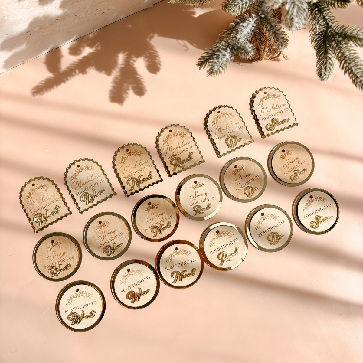 A styled flat-lay of round and arched paper gift tags with gold foil lettering, arranged on a warm beige surface with soft shadows and a decorative pine branch in the corner.