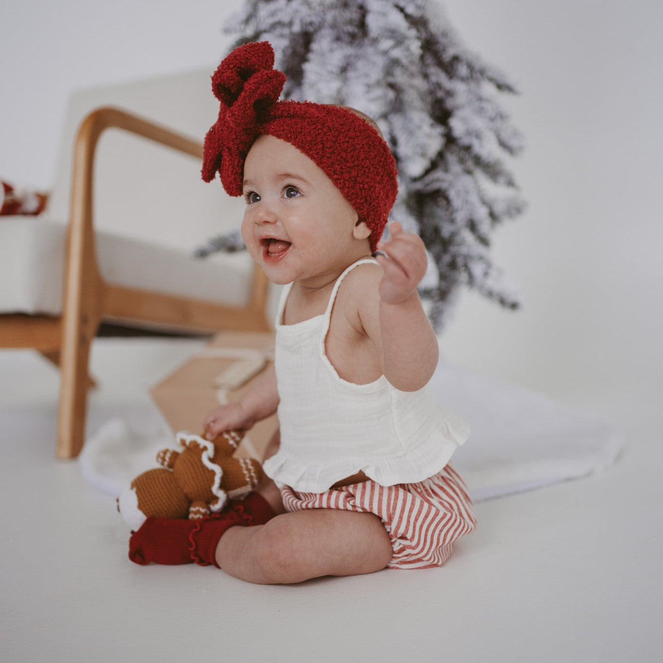 Baby in a white frill top, red stripe bloomers and a red knit bow headband beside a flocked tree