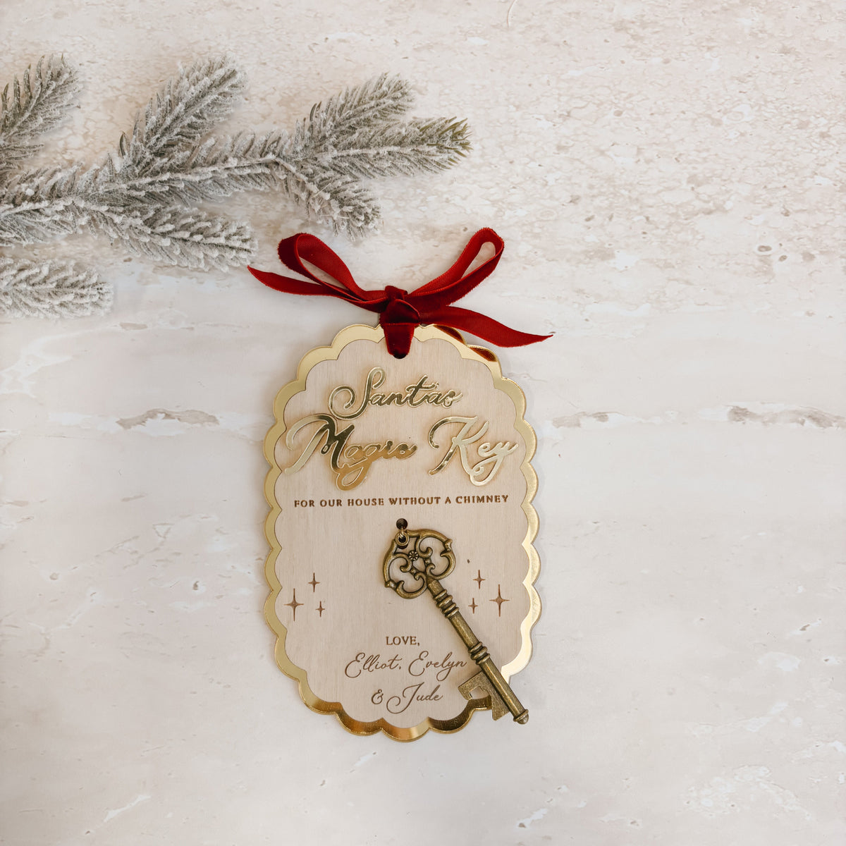A scalloped cream gift tag with gold trim, featuring an engraved vintage key and red ribbon, styled on a textured light beige surface with a silver pine branch.