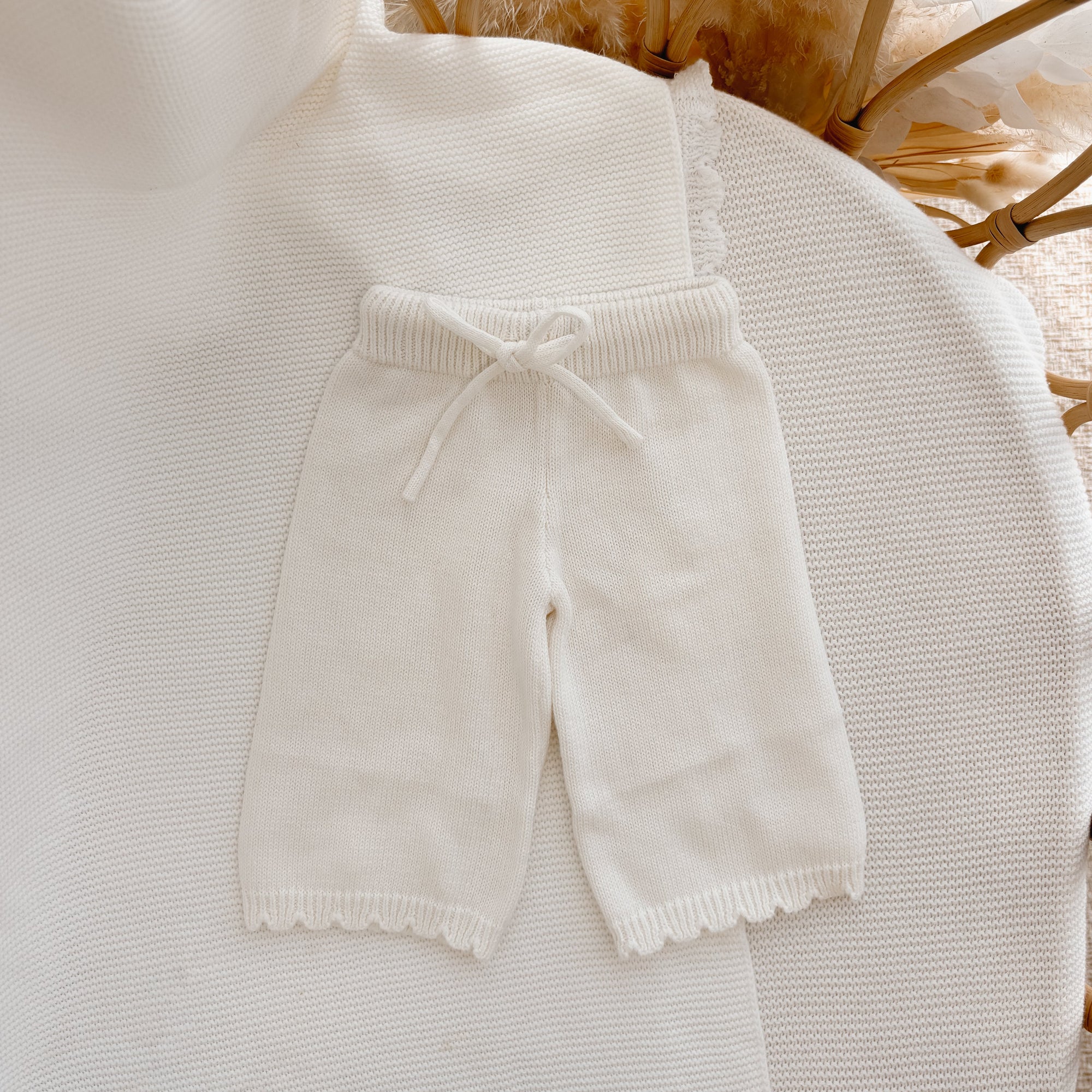 Cream knit baby pants with a drawstring waist and scalloped hem, flat lay in a bassinet