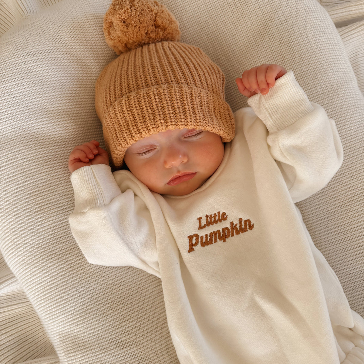Close-up of a sleeping newborn in a cream romper embroidered with Little Pumpkin in caramel script, wearing a tan ribbed pom beanie