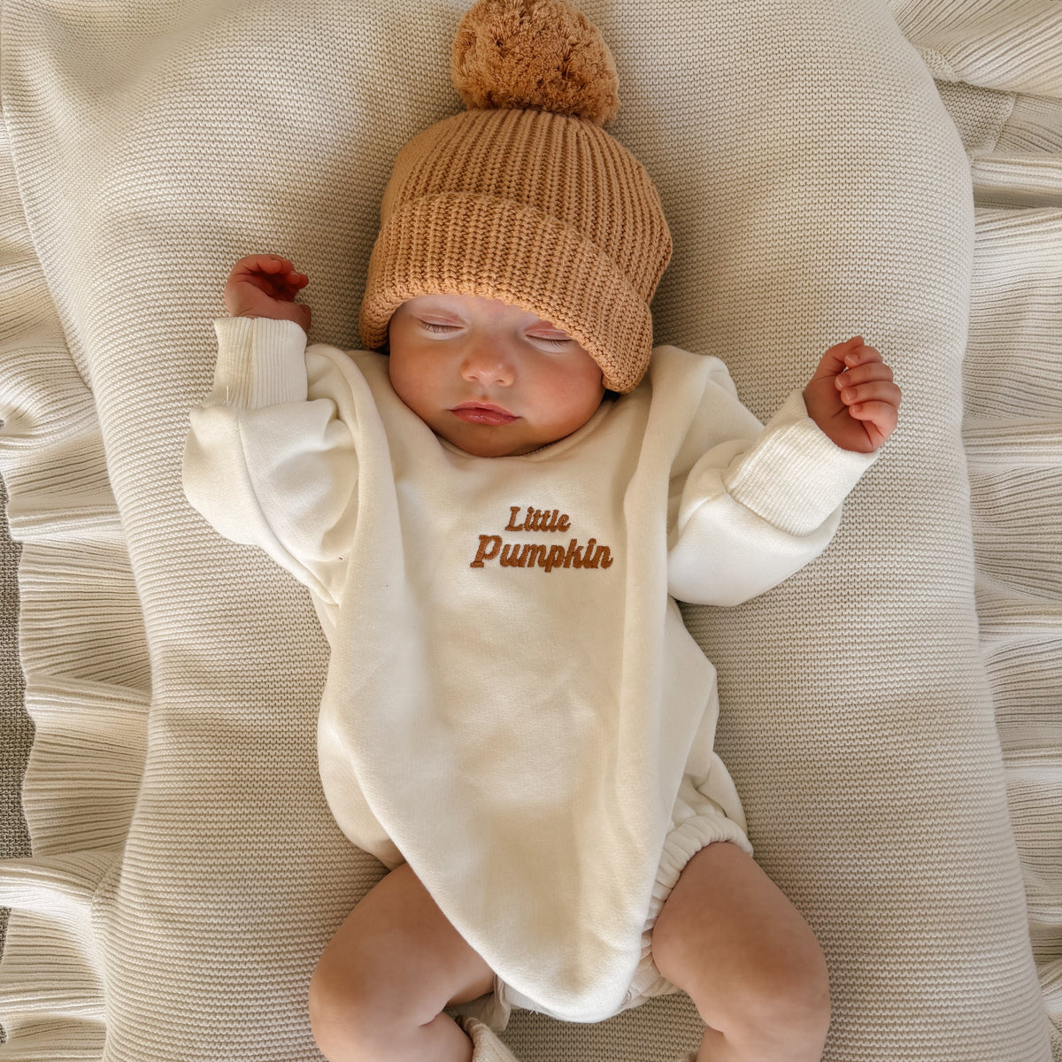 Sleeping newborn in a cream Little Pumpkin embroidered romper and tan pom beanie, arms stretched out on a ruffled knit blanket