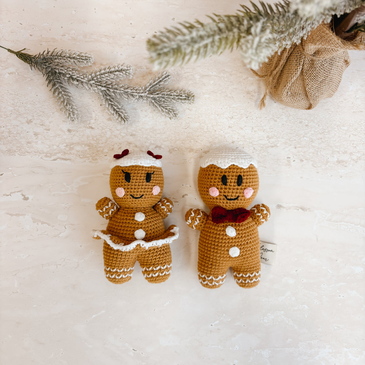 Two small crocheted gingerbread figures, one male and one female, displayed on a light textured surface with decorative snowy pine branches and a burlap sack.