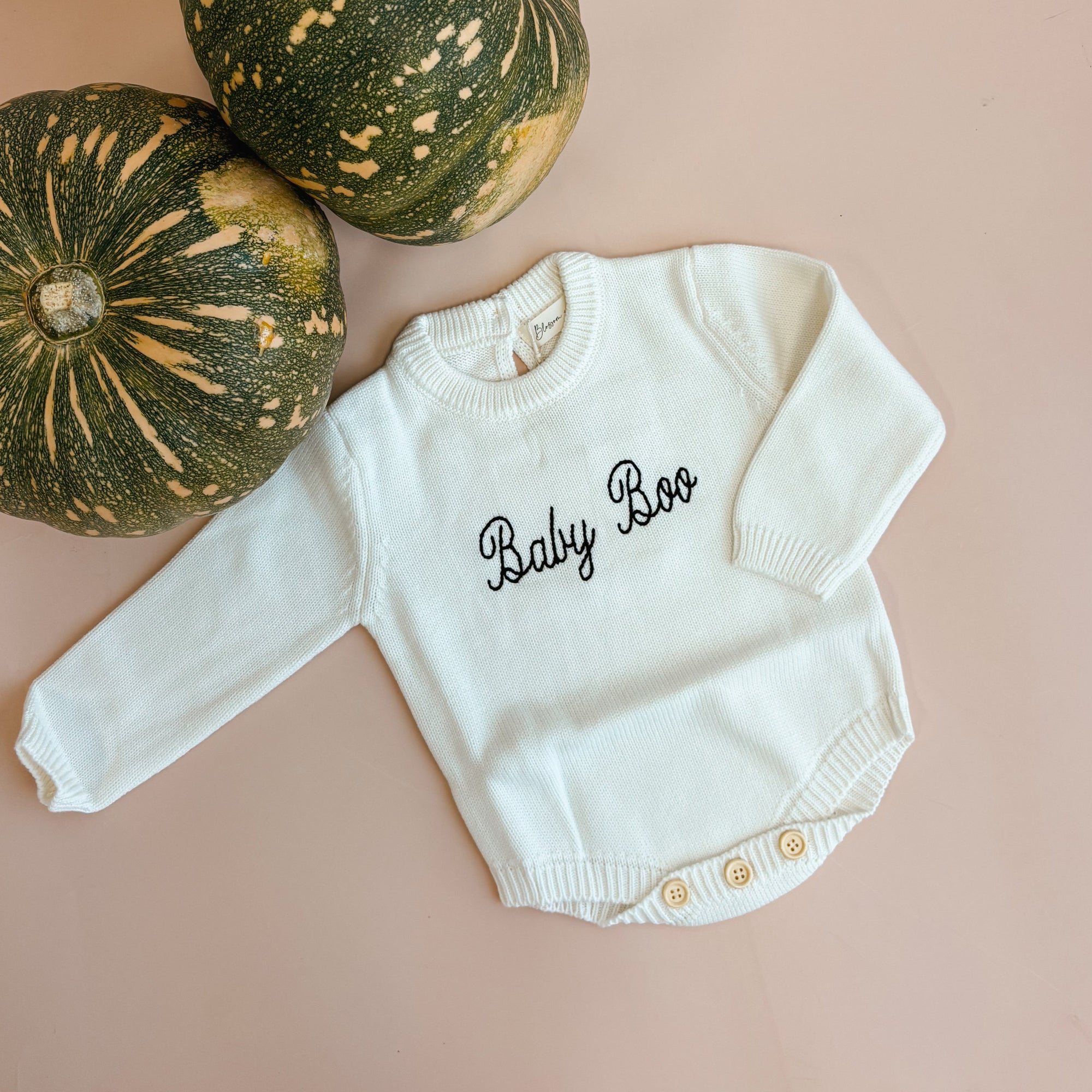 An off-white long-sleeve baby bodysuit with 'Baby Boo' embroidered in black script on the chest, styled on a dusty pink surface next to two green and yellow striped pumpkins.