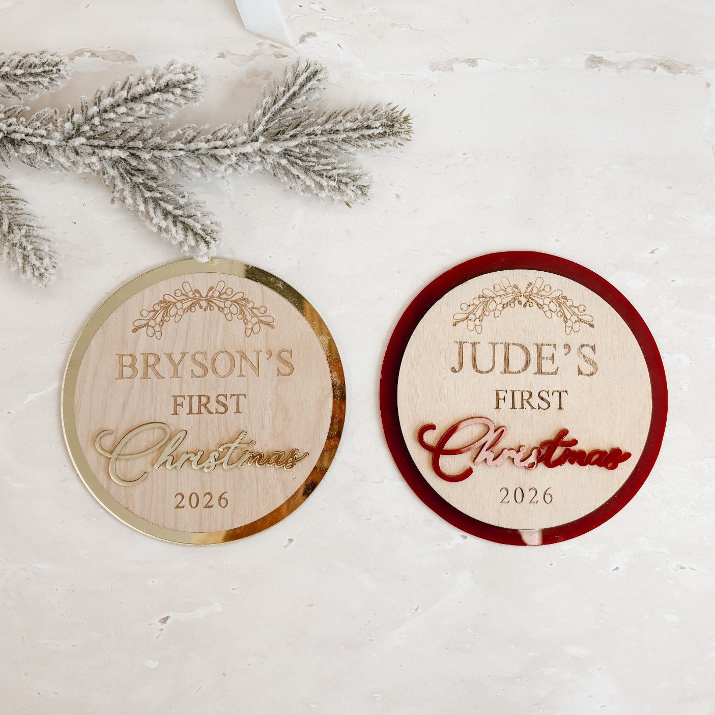 Two round personalized Christmas gift tags on a light textured surface, with a kraft gift box and white ribbon in the background and silver pine branches as props.