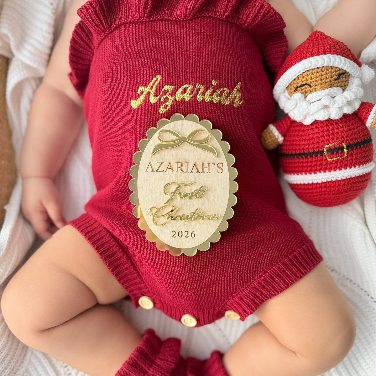A baby wearing a personalized red knit Christmas romper with gold lettering that reads 'Azariah' and a gold oval patch reading 'Azariah's First Christmas 2026'. The baby is also wearing matching red knit booties and a red knit Santa hat.