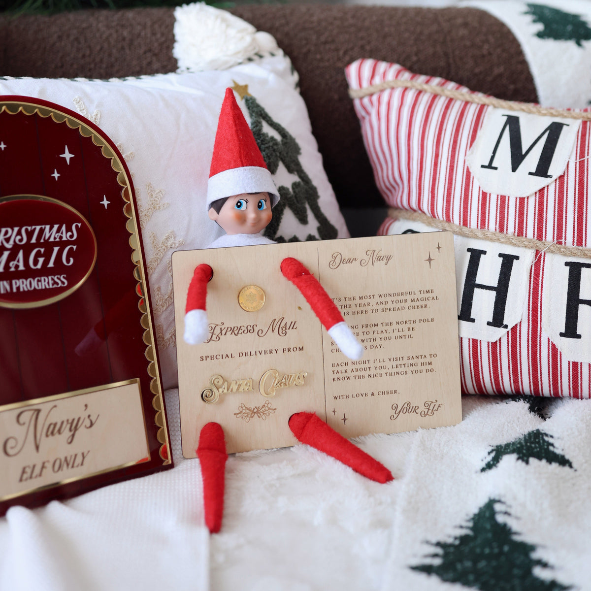 A Christmas-themed lifestyle scene featuring an Elf on the Shelf toy sitting on a patterned throw blanket next to a personalized letter card, red and white pillows, and a Christmas sign.