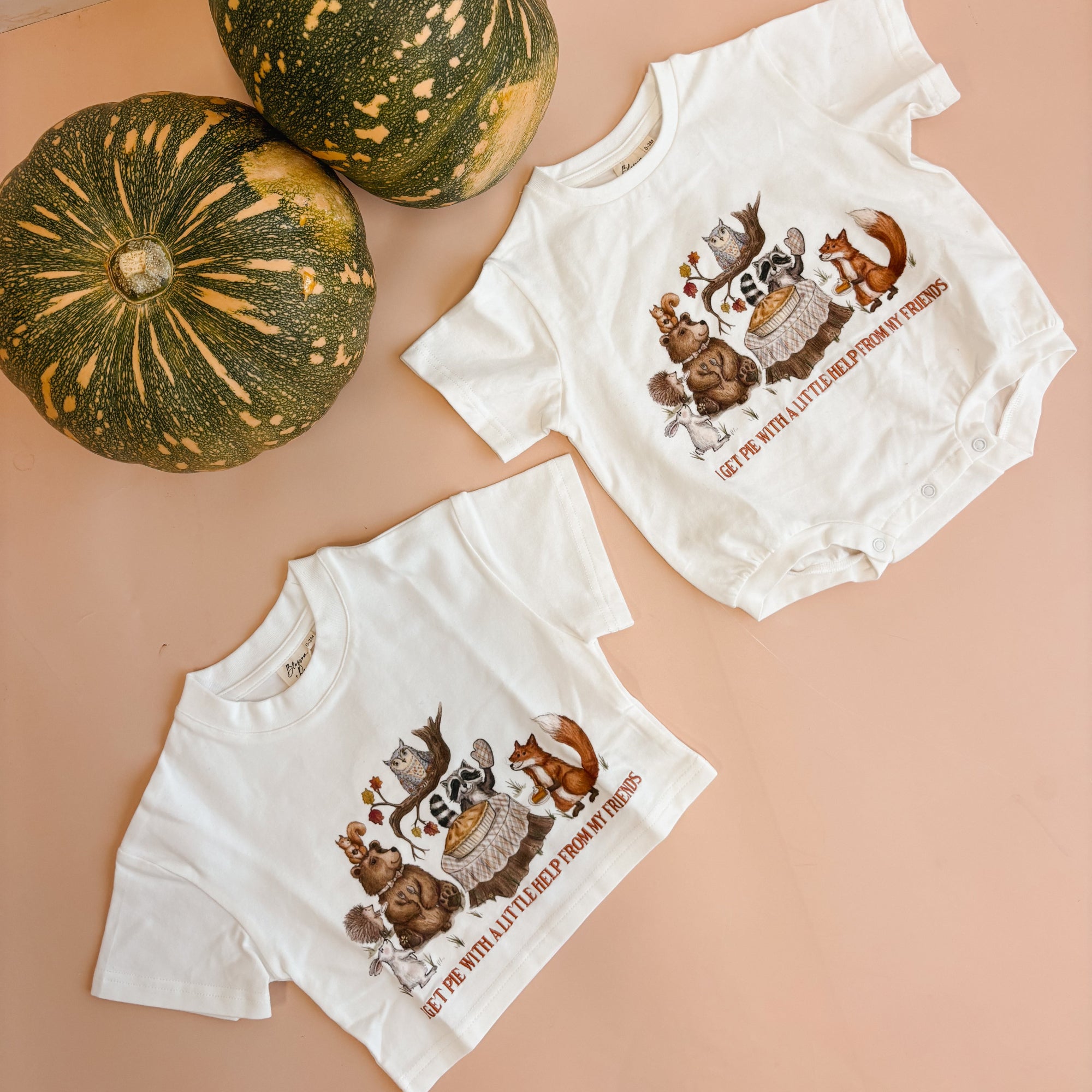 Two white short-sleeve baby bodysuits with a fall-themed woodland animal graphic and text, arranged on a peach-colored surface next to two green pumpkins.