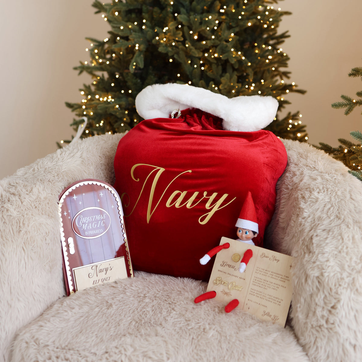 Personalised burgundy velvet Santa sack embroidered with the name Navy on a cream armchair with a Christmas Eve box and elf