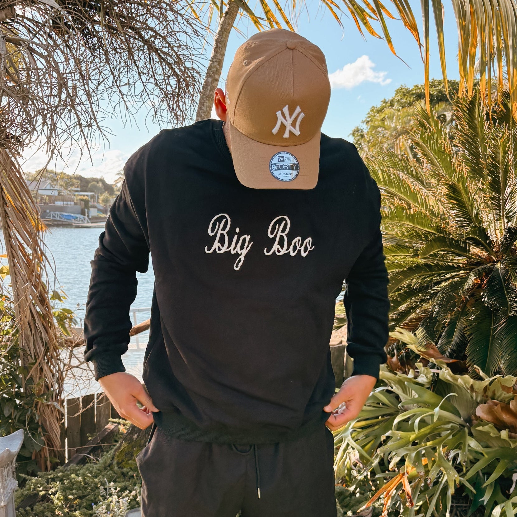 A man wearing a black long-sleeve sweatshirt with 'Big Boo' written on the front, black shorts, standing outdoors in a tropical garden setting.