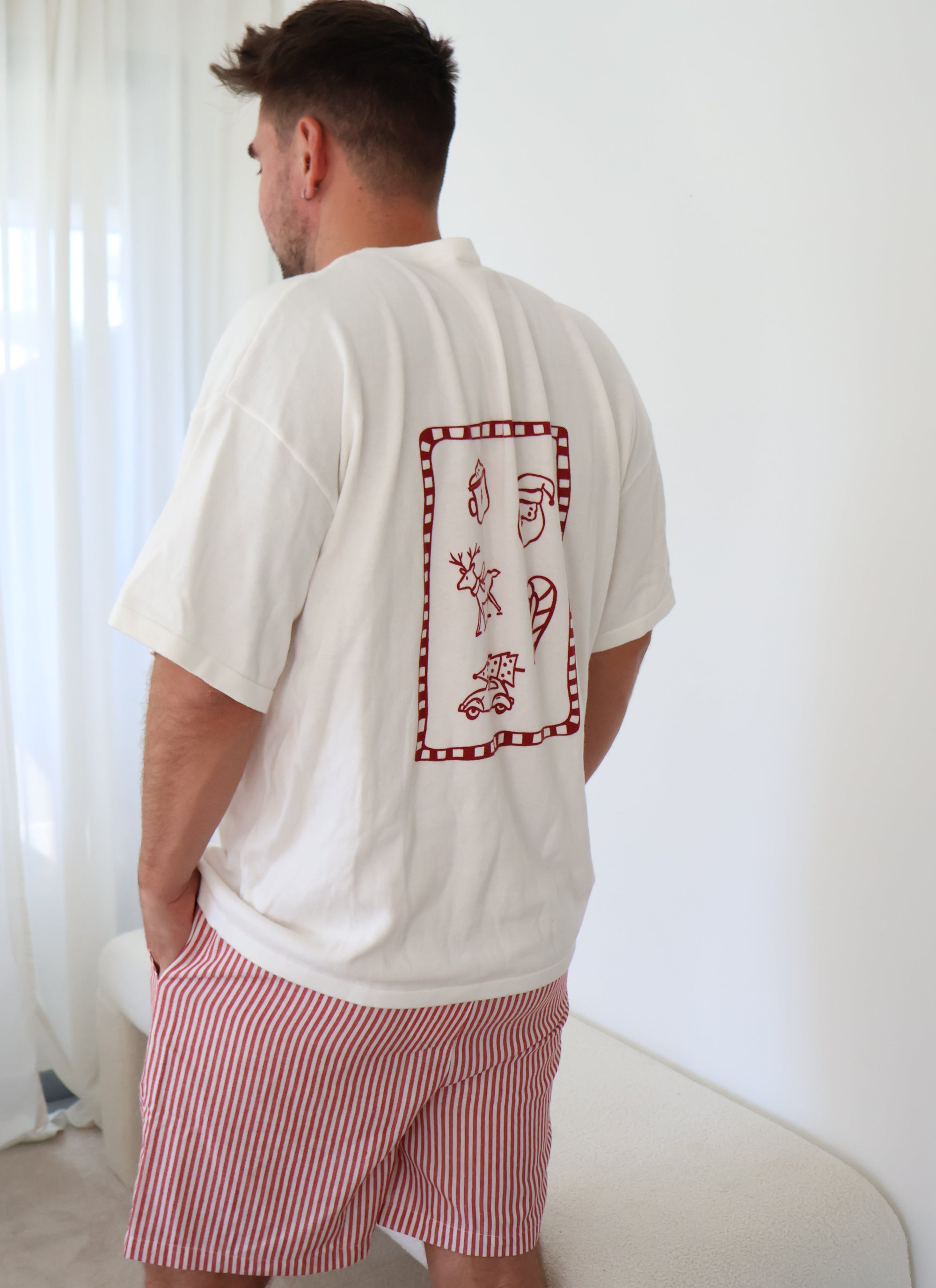 Back view of a men's white Christmas tee with a red doodle print of a reindeer, stocking and Santa