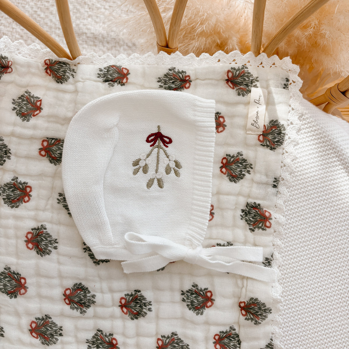 White knit baby bonnet embroidered with mistletoe and a red bow, on a wreath print muslin blanket