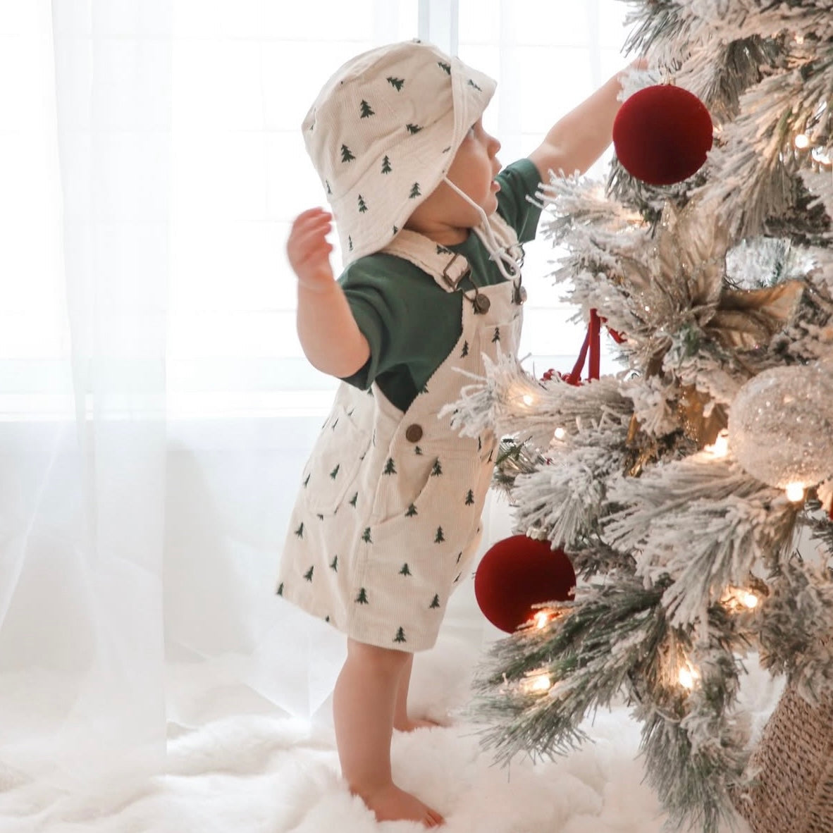 Toddler in Christmas tree print corduroy overalls and bucket hat reaching for a bauble on a flocked tree
