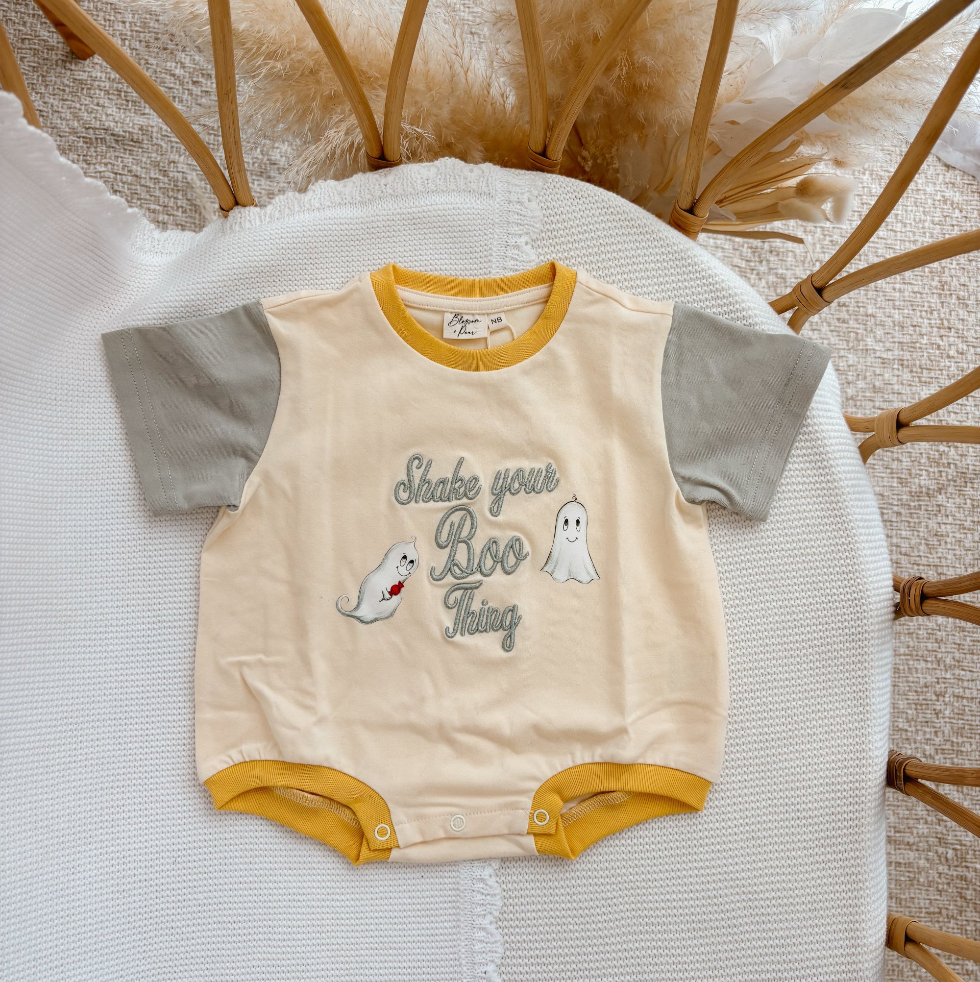 A baby short-sleeve bodysuit laid flat on a white textured blanket, surrounded by a wicker chair and pampas grass. The bodysuit has a cream body, mustard yellow trim, and gray sleeves, featuring a 'Shake your Boo Thing' graphic with ghost illustrations.