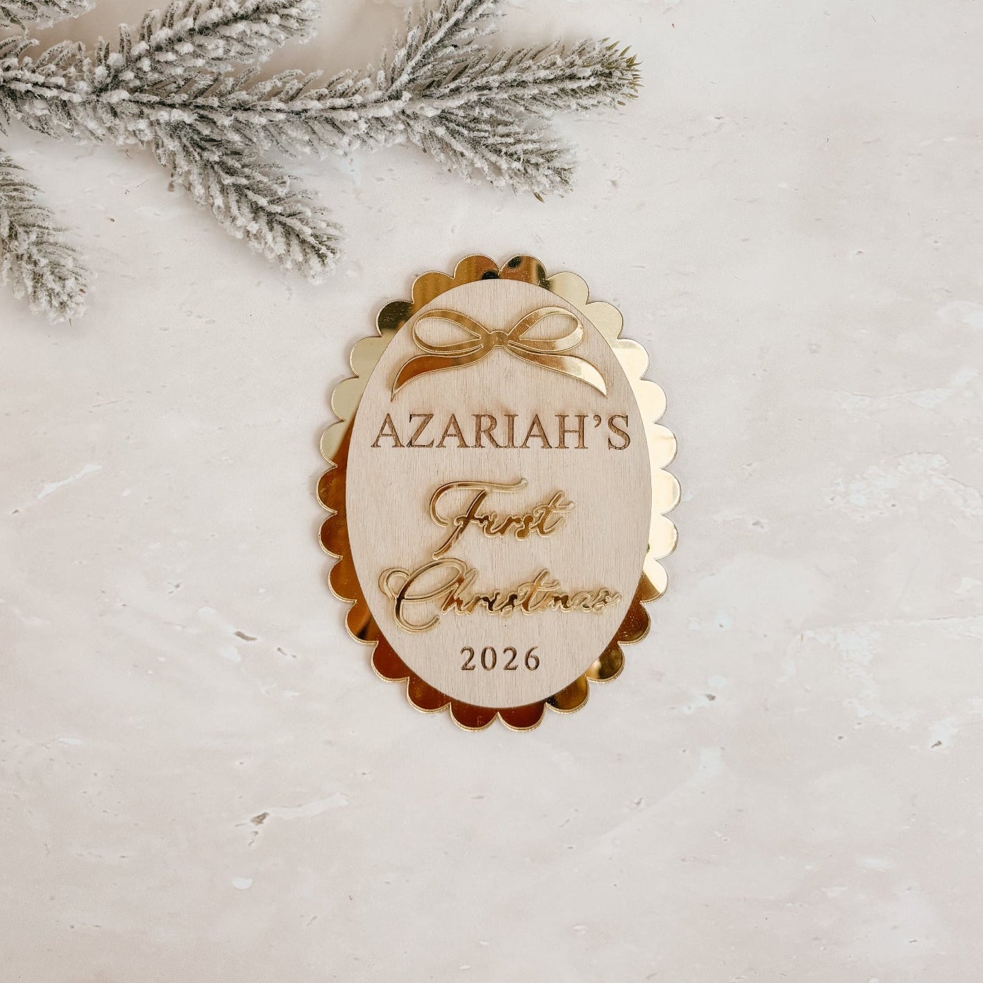 A personalized oval Christmas ornament with scalloped edges and gold foil lettering that reads 'AZARIAH'S First Christmas 2026', styled on a light textured surface with a kraft gift box, white ribbon, and silver pine branches.