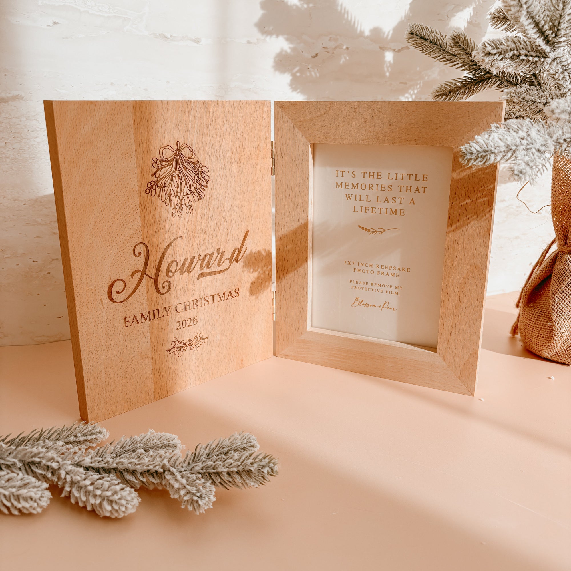 Personalised wooden keepsake photo frame standing upright, engraved 'Howard Family Christmas 2026' with mistletoe detail, frosted pine branch beside it
