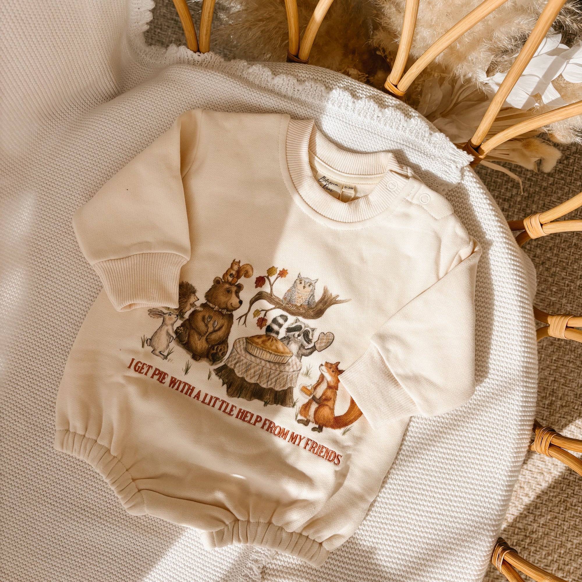 A cream-colored baby sweatshirt with a graphic of cartoon animals and the text 'I GET PIE WITH A LITTLE HELP FROM MY FRIENDS', laid flat on a textured white blanket with wicker and faux fur props.