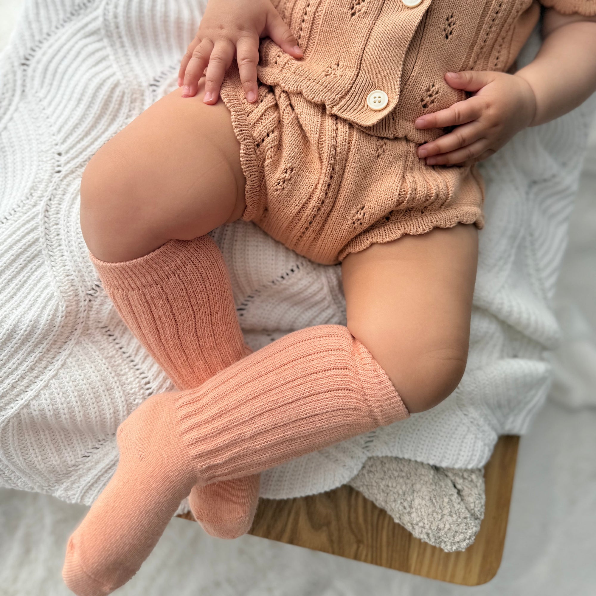 Baby wearing a tan pointelle knit romper and matching apricot knee high socks on a white knit blanket