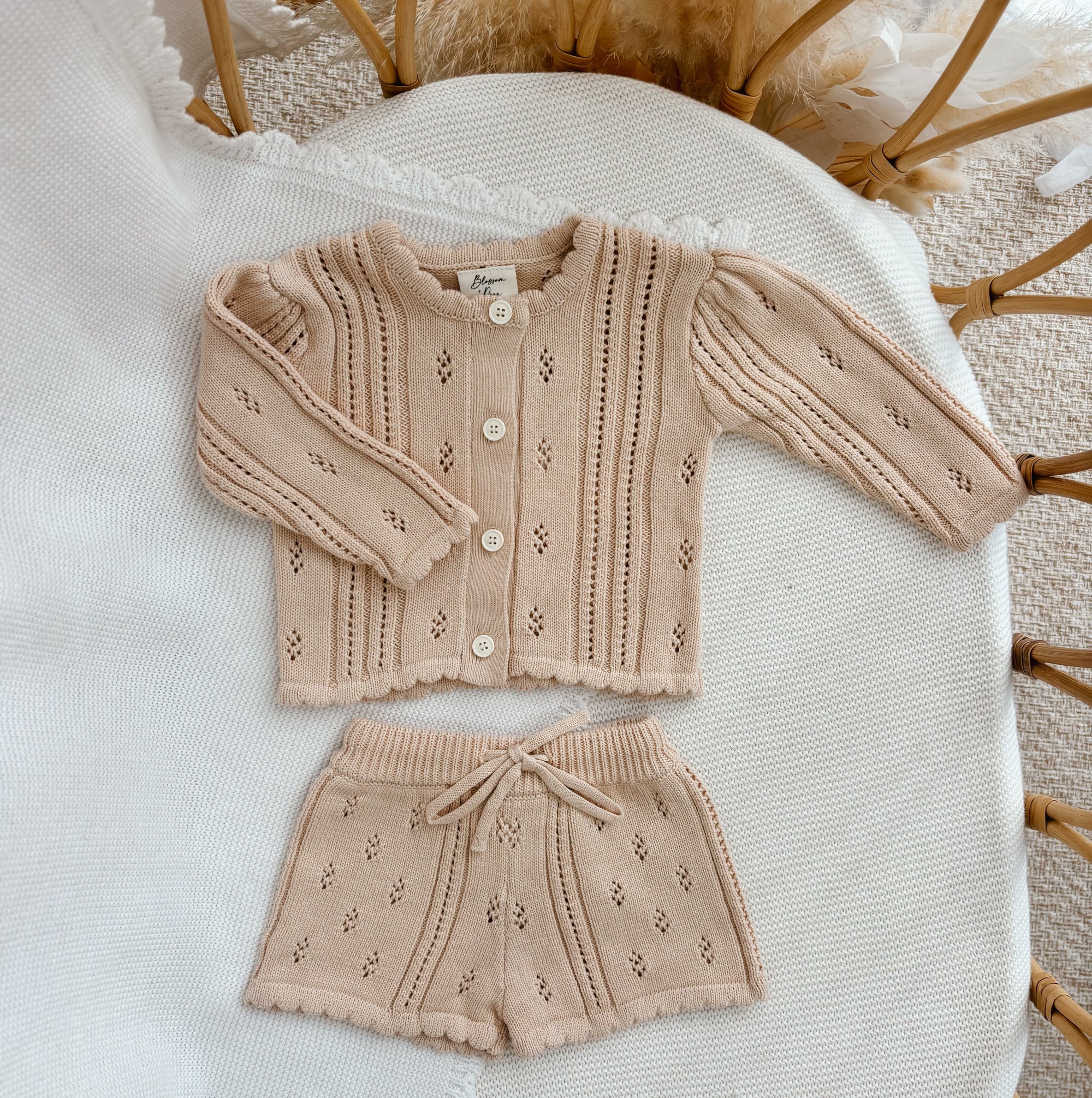Oat pointelle knit baby cardigan and drawstring shorts set, flat lay on a cream blanket in a bassinet