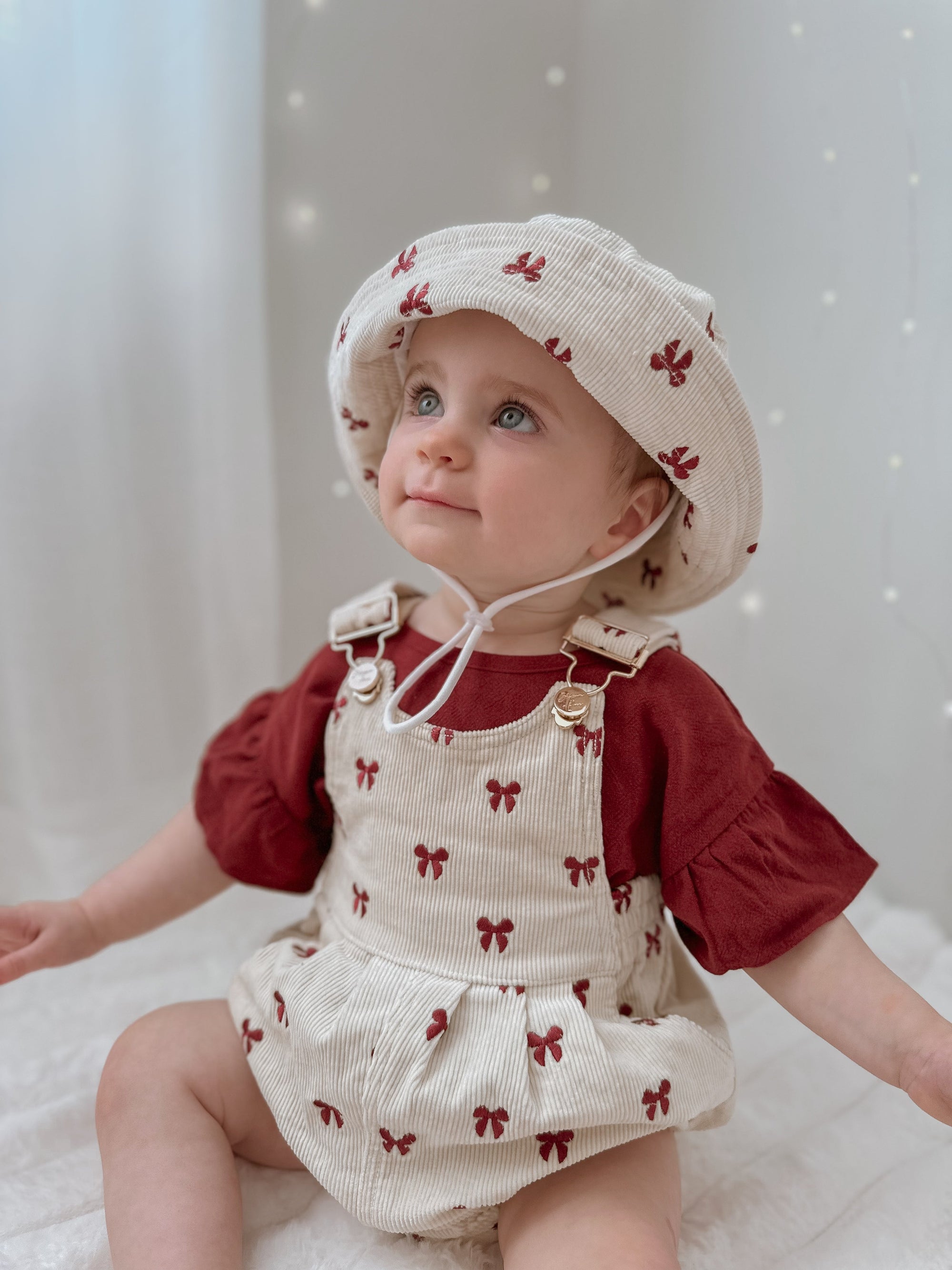 Baby in a red bow print corduroy overall romper, matching bucket hat and red puff sleeve tee