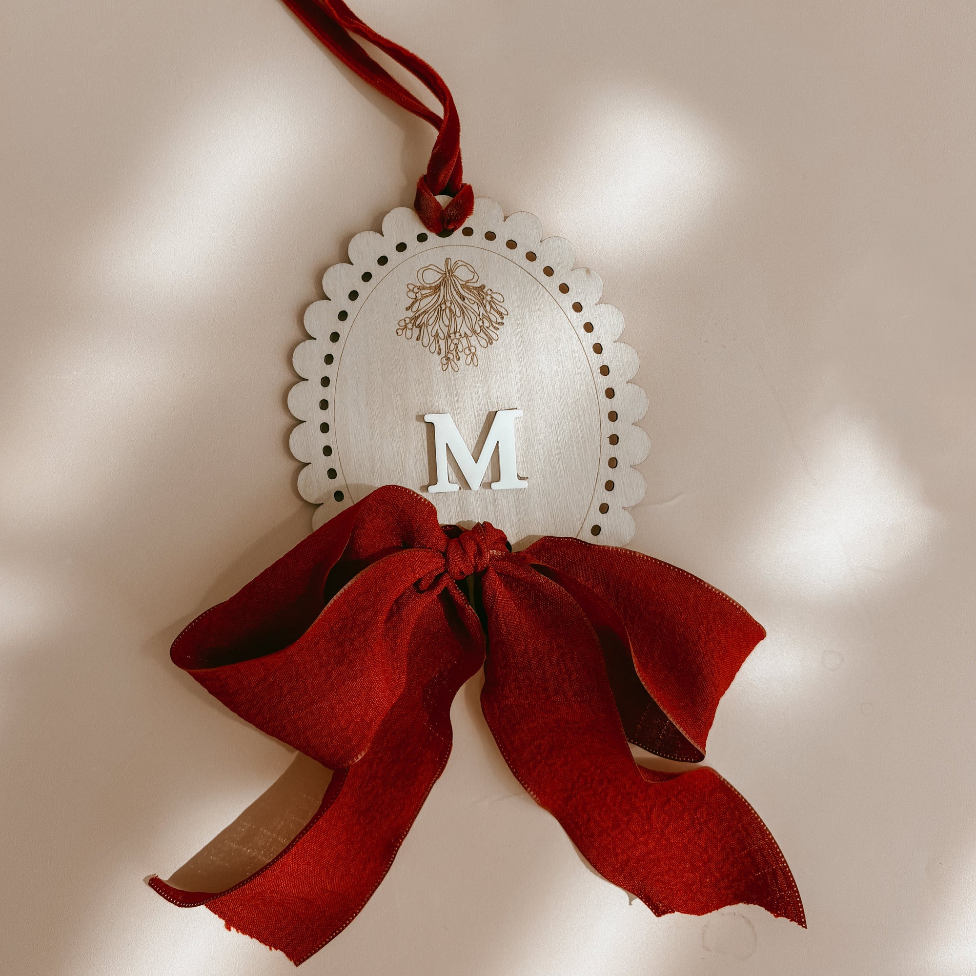 A personalized oval wooden ornament with a scalloped edge, featuring a cutout letter 'M' and a small pine tree design, tied with a red ribbon and bow against a warm beige background with soft leaf shadows.