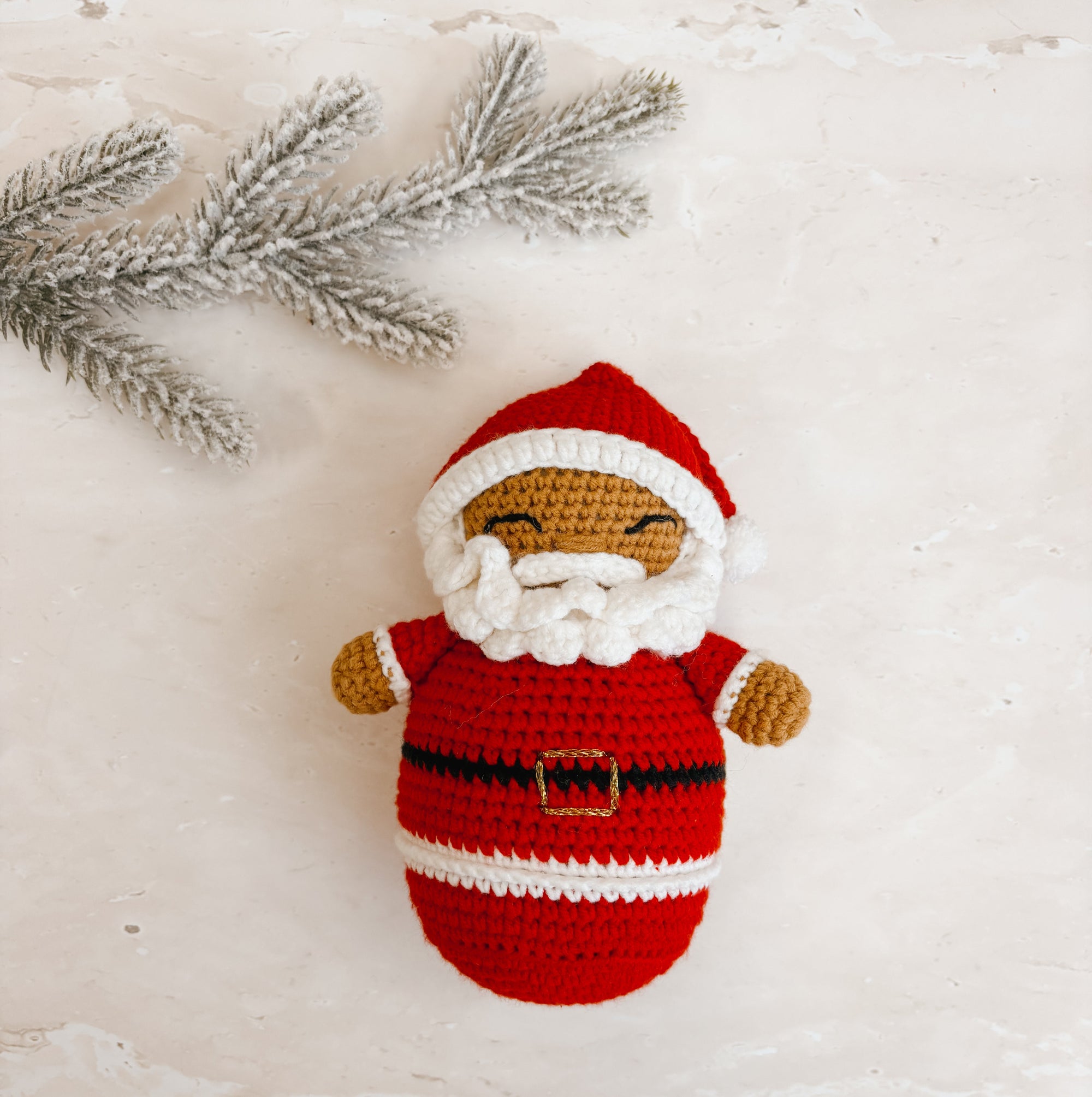 A crocheted Santa Claus doll with a red and white outfit, white beard, and brown face, styled on a light textured surface with a silver pine branch in the corner.