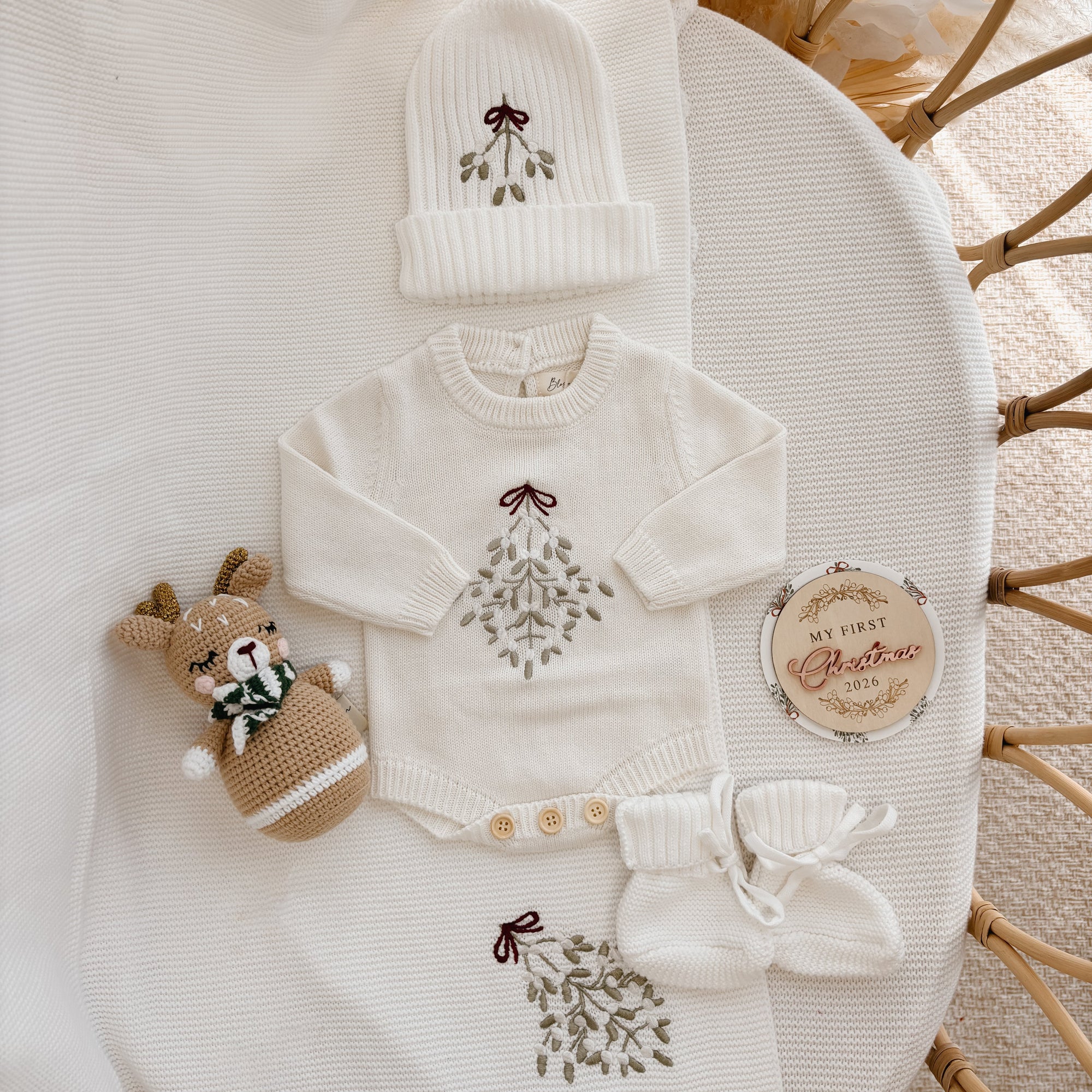 Mistletoe embroidered knit baby romper and beanie flat lay with a crochet reindeer rattle and milestone disc