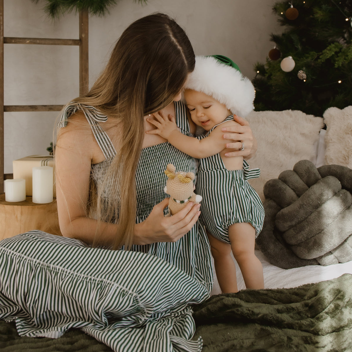 A mother and child wearing matching green and white striped holiday outfits, sitting together on a bed in a cozy, decorated Christmas room. The child is wearing a white Santa hat, and the background features a Christmas tree, wreath, and candles.