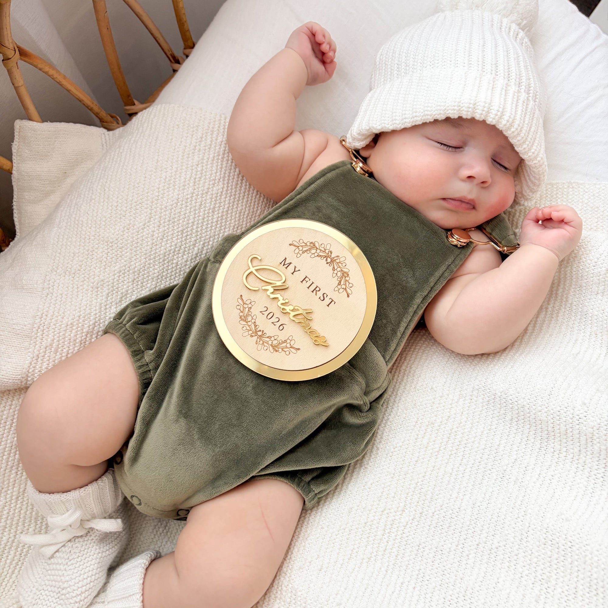 Sleeping baby in an olive velvet overall romper with a gold My First Christmas 2026 keepsake disc resting on the chest