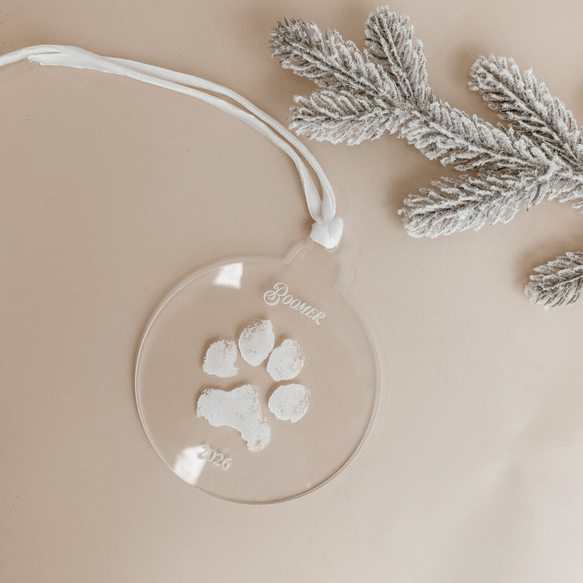 Clear acrylic pet paw print Christmas ornament personalised with the pet's name and 2026, on a white ribbon beside a frosted pine branch