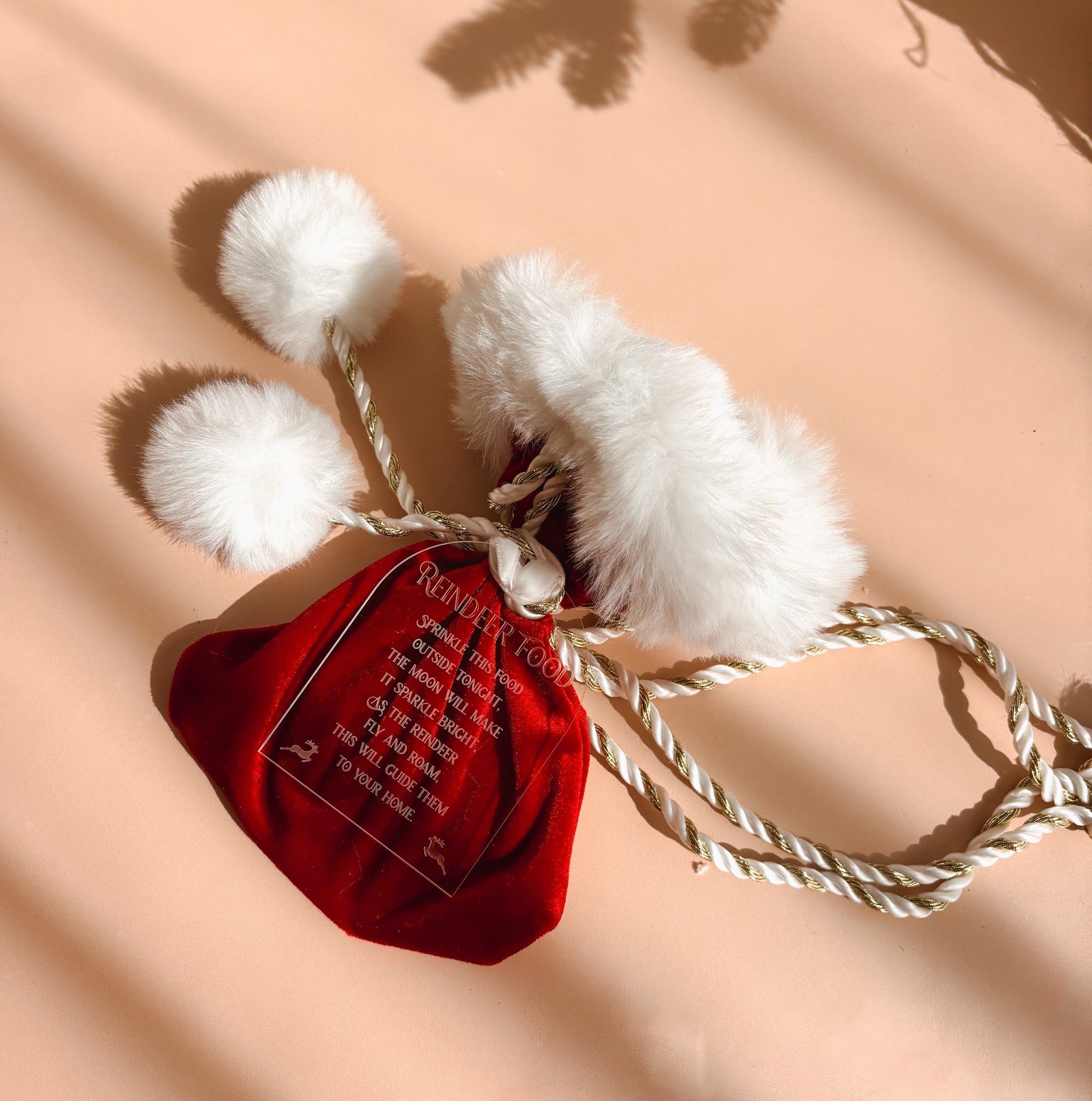 A red velvet drawstring pouch with white faux fur trim and pom-poms, attached to a twisted rope, laid on a peach background with leaf shadows.