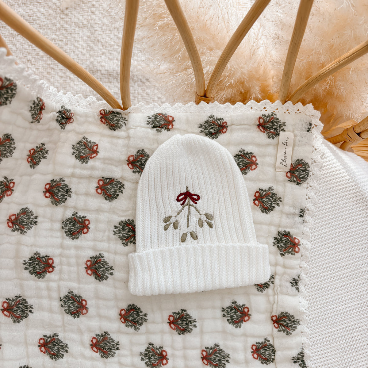 White ribbed knit baby beanie embroidered with mistletoe and a red bow on a wreath print blanket