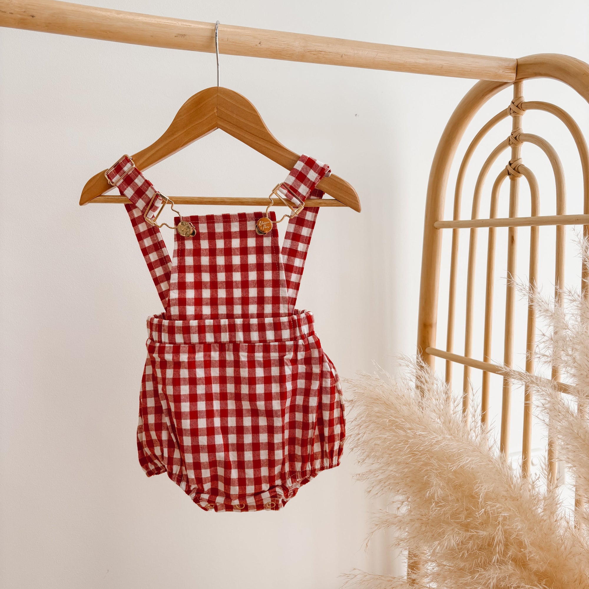 Red gingham baby overall romper with adjustable straps, hanging on a wooden hanger