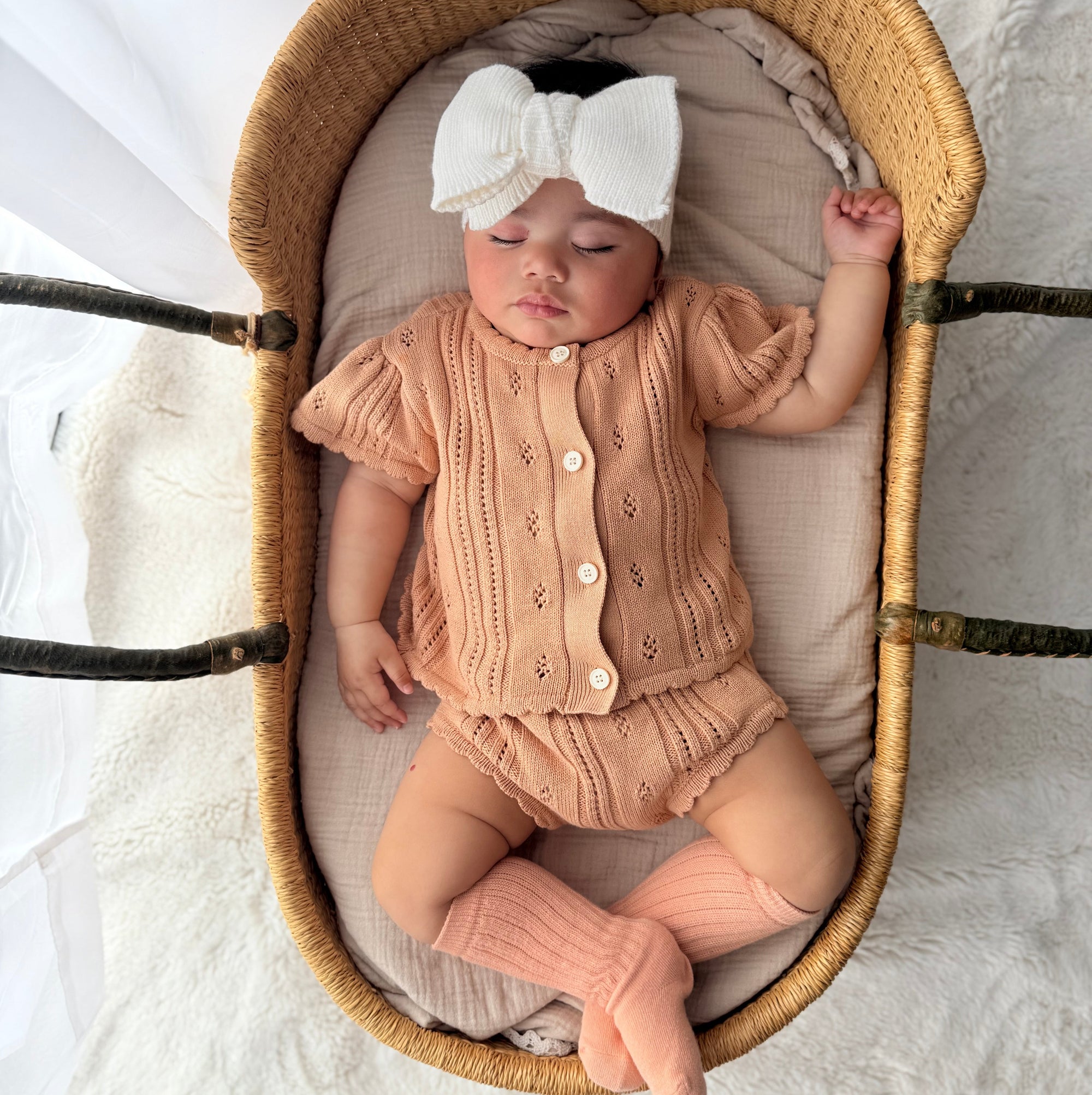 Sleeping baby in an apricot pointelle knit set and white bow headband lying in a woven bassinet