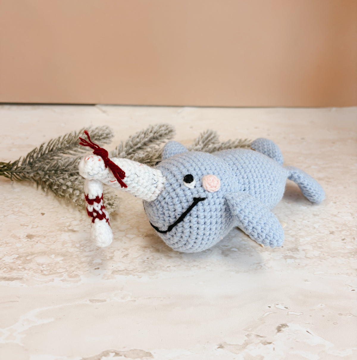 A light blue crocheted narwhal plush toy lying on a textured beige surface next to a small white and red crocheted figure, with a soft beige background.