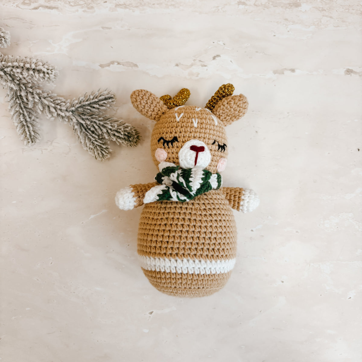 A crocheted plush reindeer doll with brown and beige yarn, a green and white striped scarf, and white accents, styled on a light beige textured surface next to a decorative pine branch.