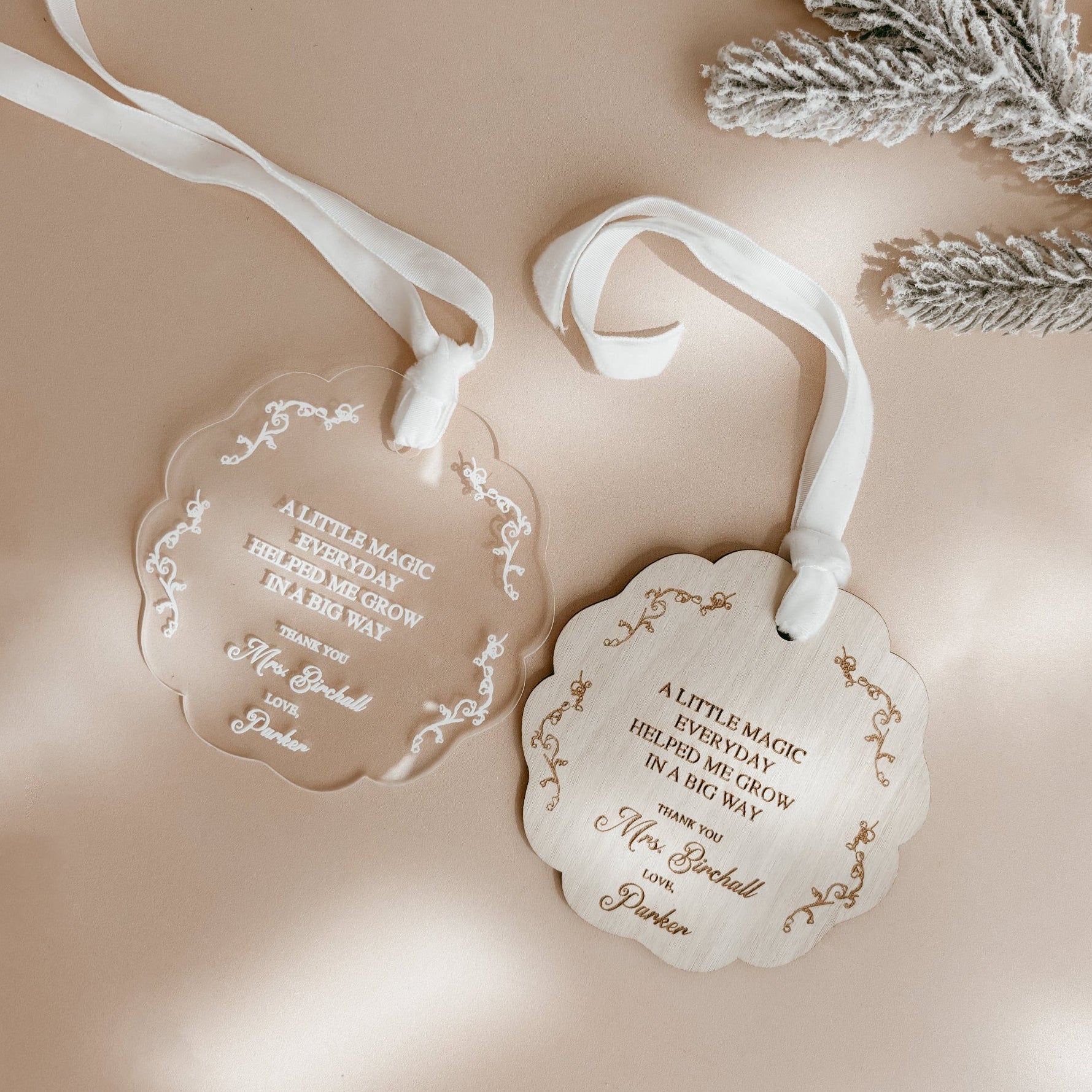 Two scalloped acrylic Christmas ornaments with white ribbons, one clear and one white, printed with gold and black text, styled on a beige background with a silver pine branch.