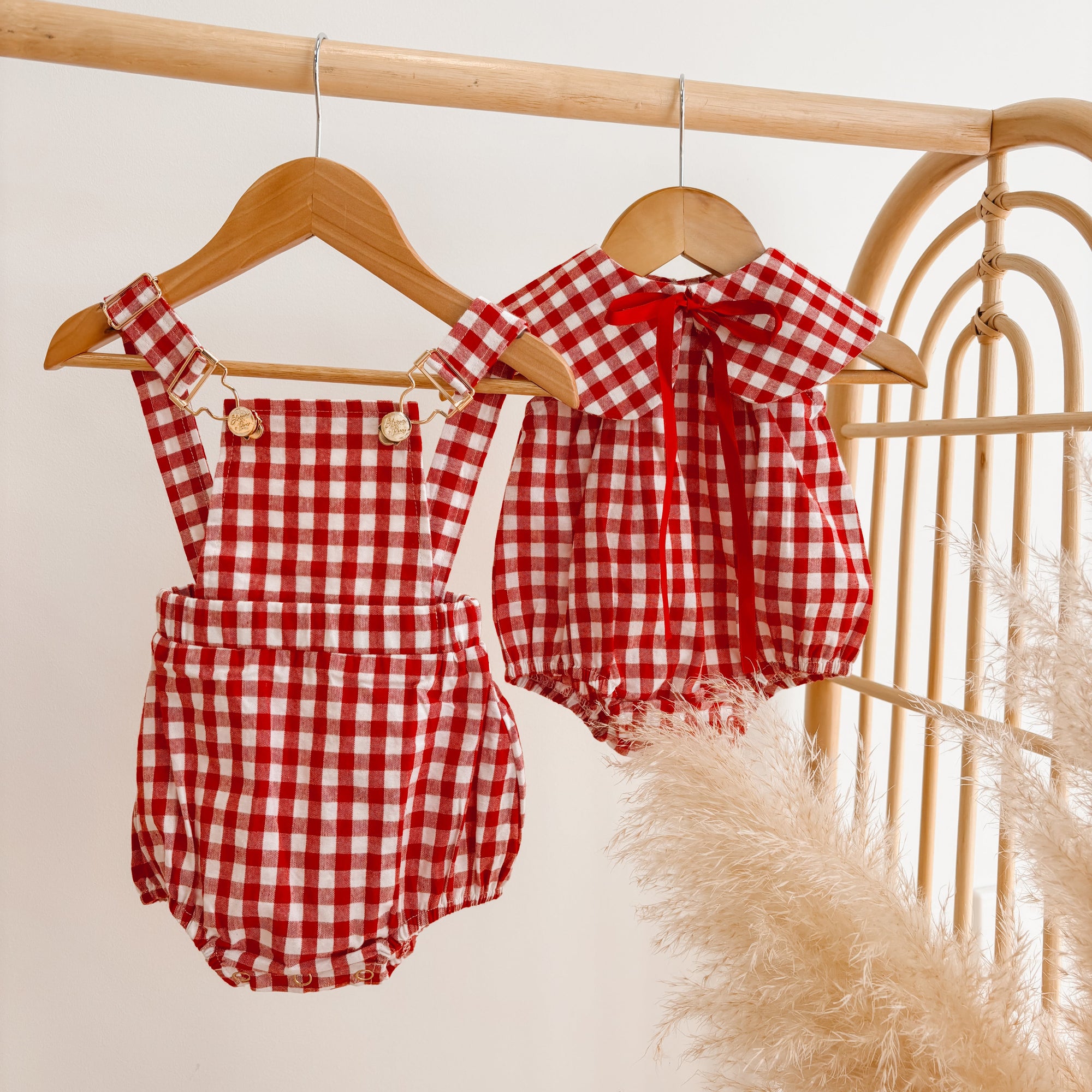 Red gingham baby overall romper and collared romper hanging side by side on a rattan rail