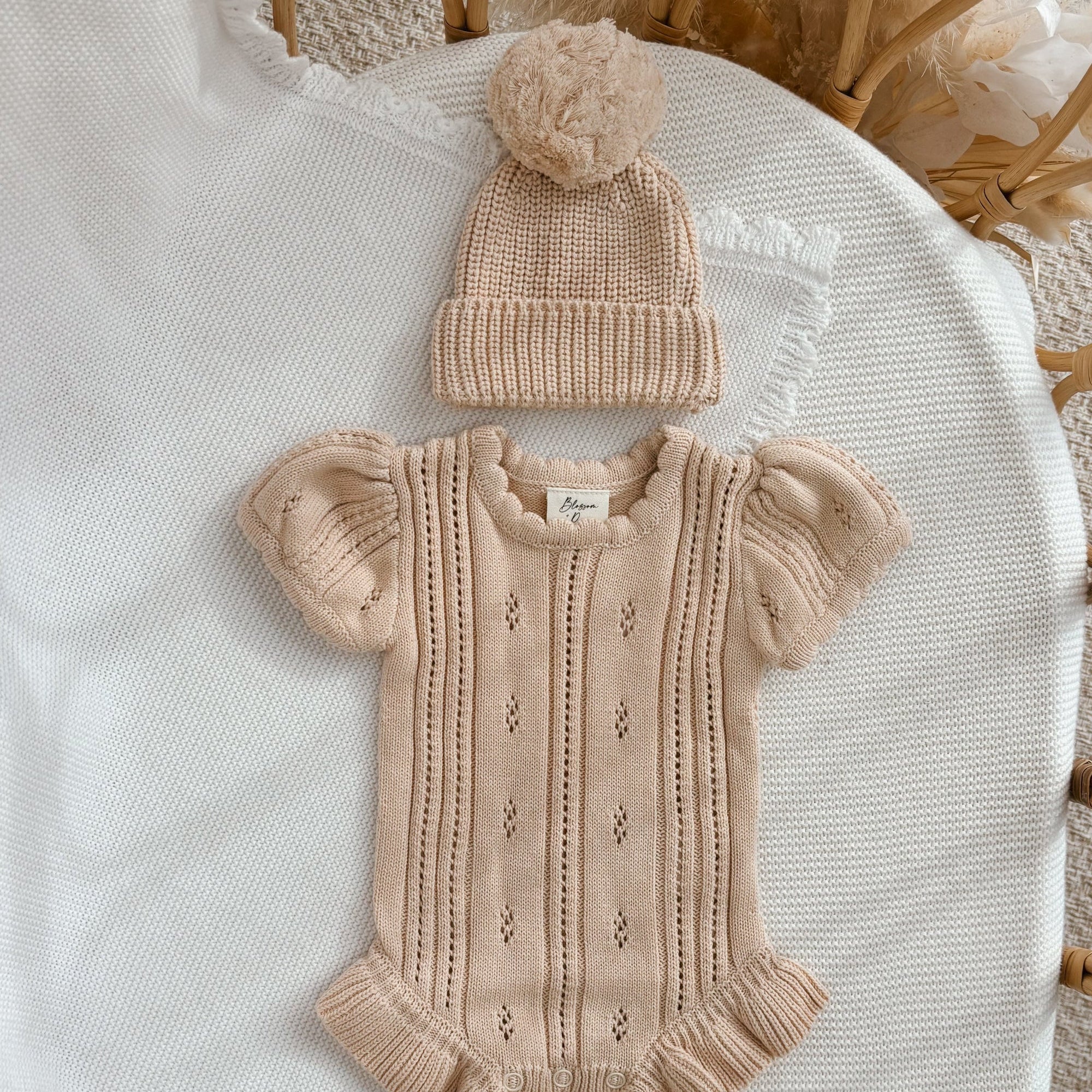 Vanilla Oat Margot Knit Romper with a matching pom beanie and white knit booties on a cream blanket