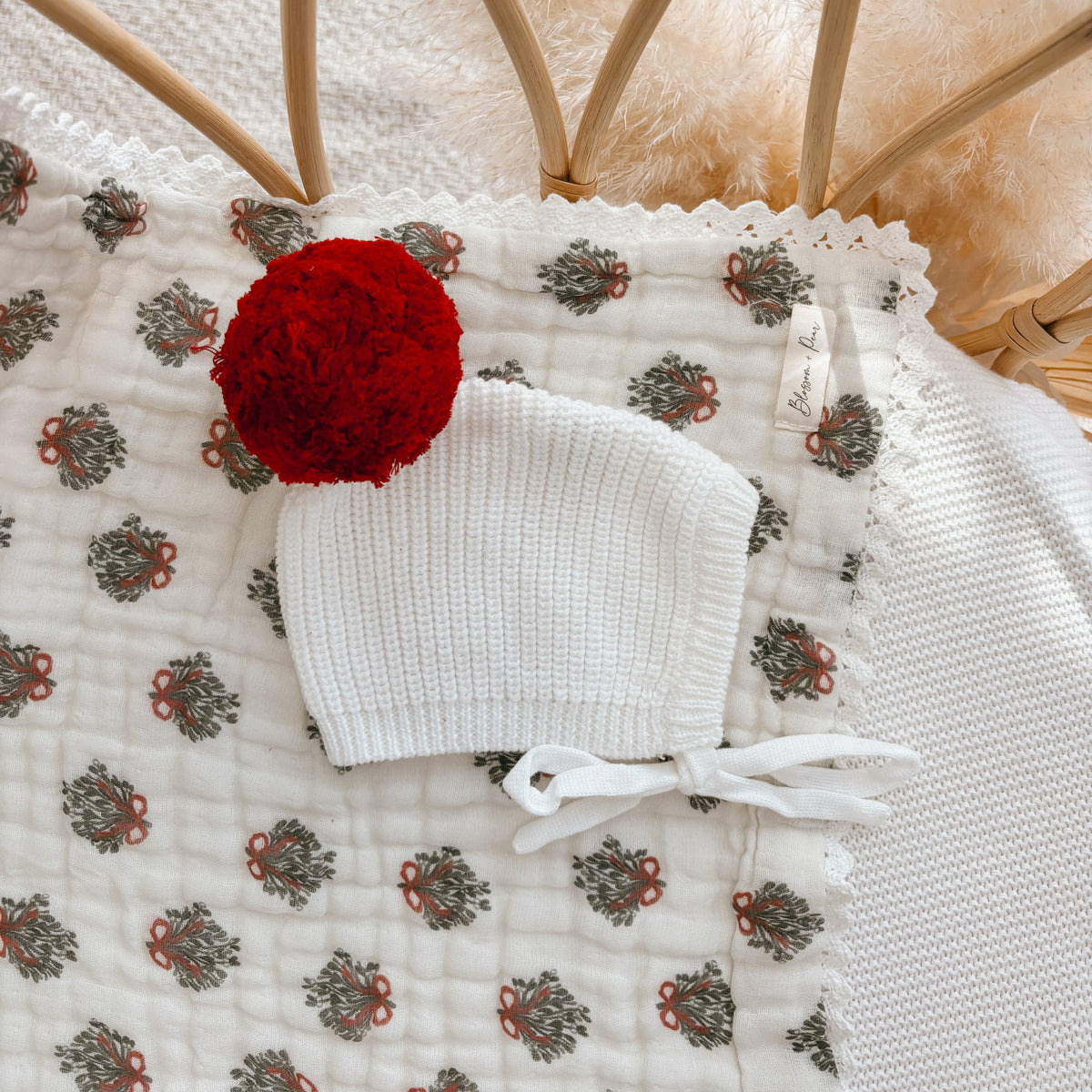 White knit baby bonnet with a red pom pom and tie ribbons on a wreath print muslin blanket