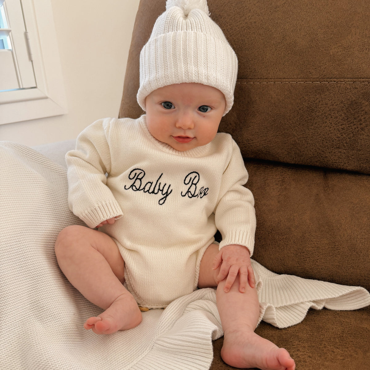 Baby in a cream knit romper hand embroidered with 'Baby Boo' and a matching white pom beanie, sitting on a lounge with a knit blanket