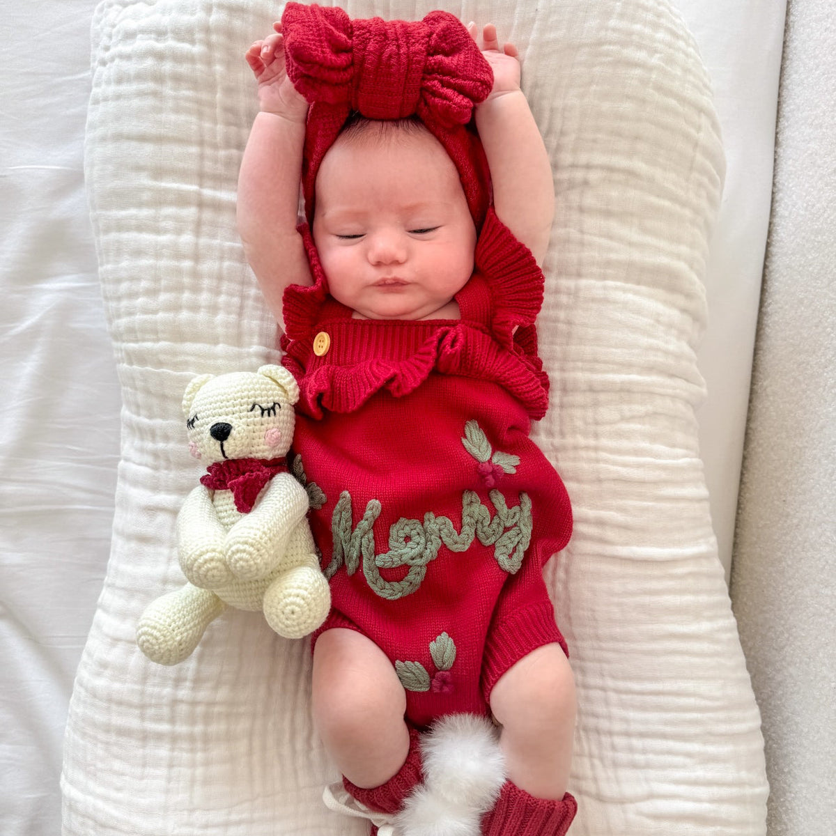 A sleeping newborn baby wearing a festive red knit holiday outfit with a large matching bow headband, red booties with white pom-poms, and holding a cream-colored crochet teddy bear. The baby is lying on a textured white pillow.