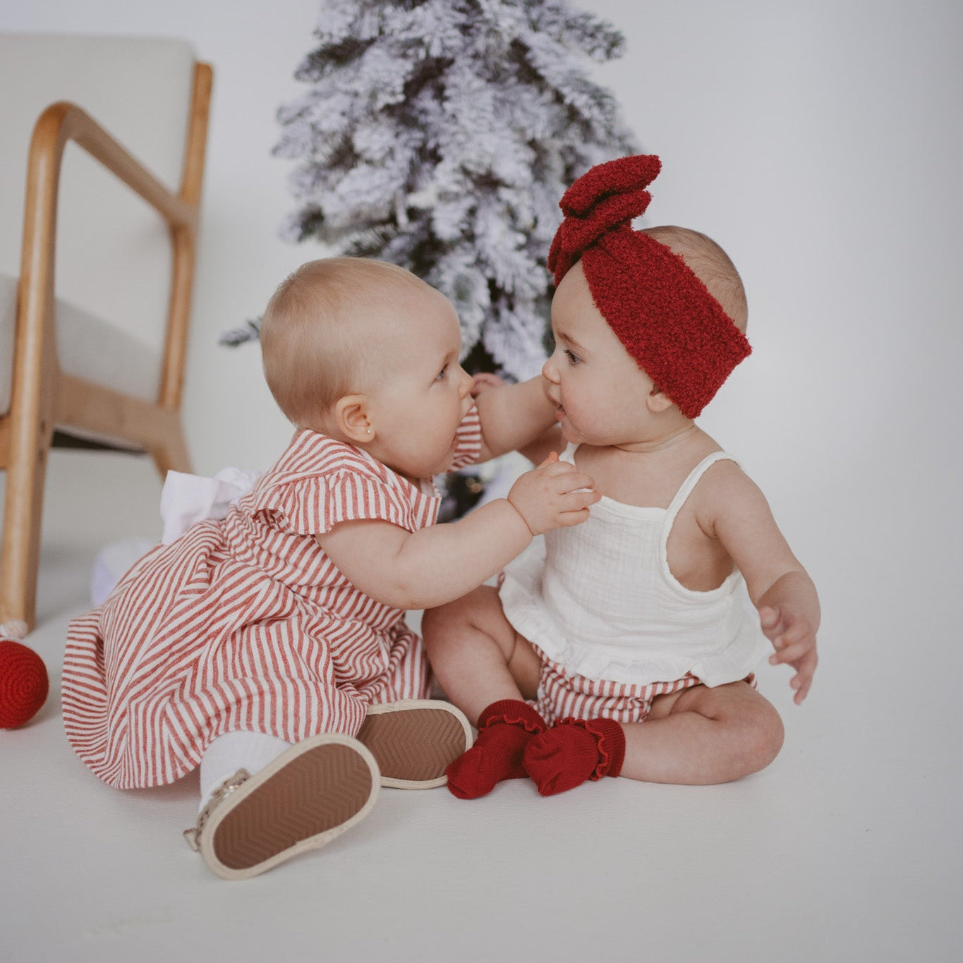 Two babies in red stripe Christmas outfits sitting together beside a flocked Christmas tree